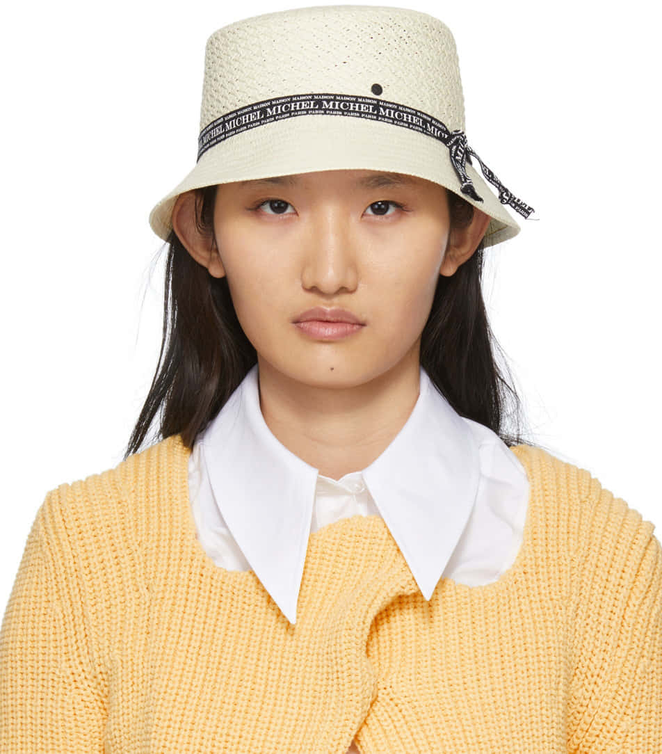 Clothes 桌布 壁紙 — 一位女士戴著一頂奶油色編織漁夫帽，上面有重複的黑色MICHEL品牌標誌，並在白色有領襯衫外穿一件黃色針織毛衣。