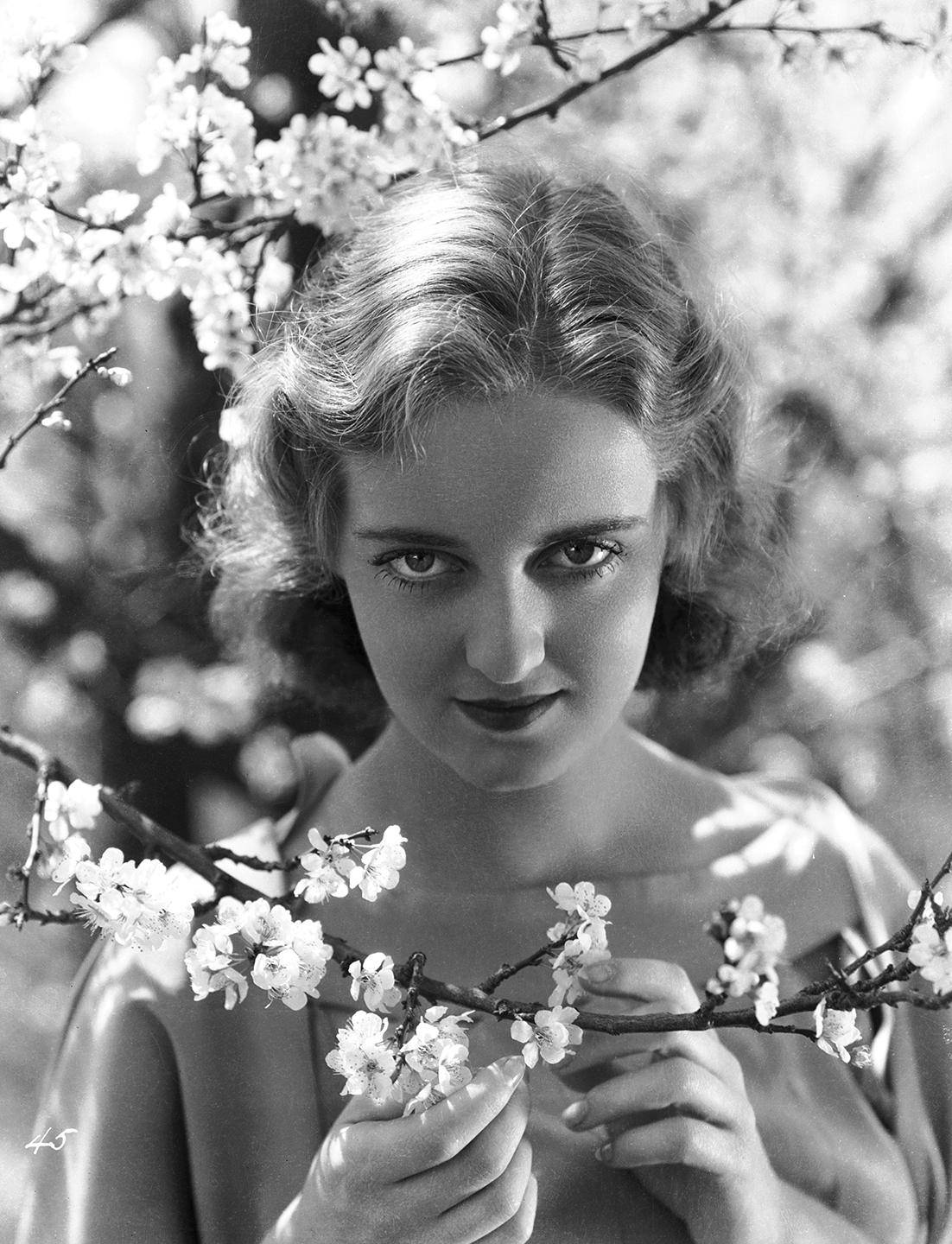 Bette Davis 桌布 壁紙 — 一位女士手持盛開的樹枝，精緻的花朵框住她的臉龐的復古黑白肖像。