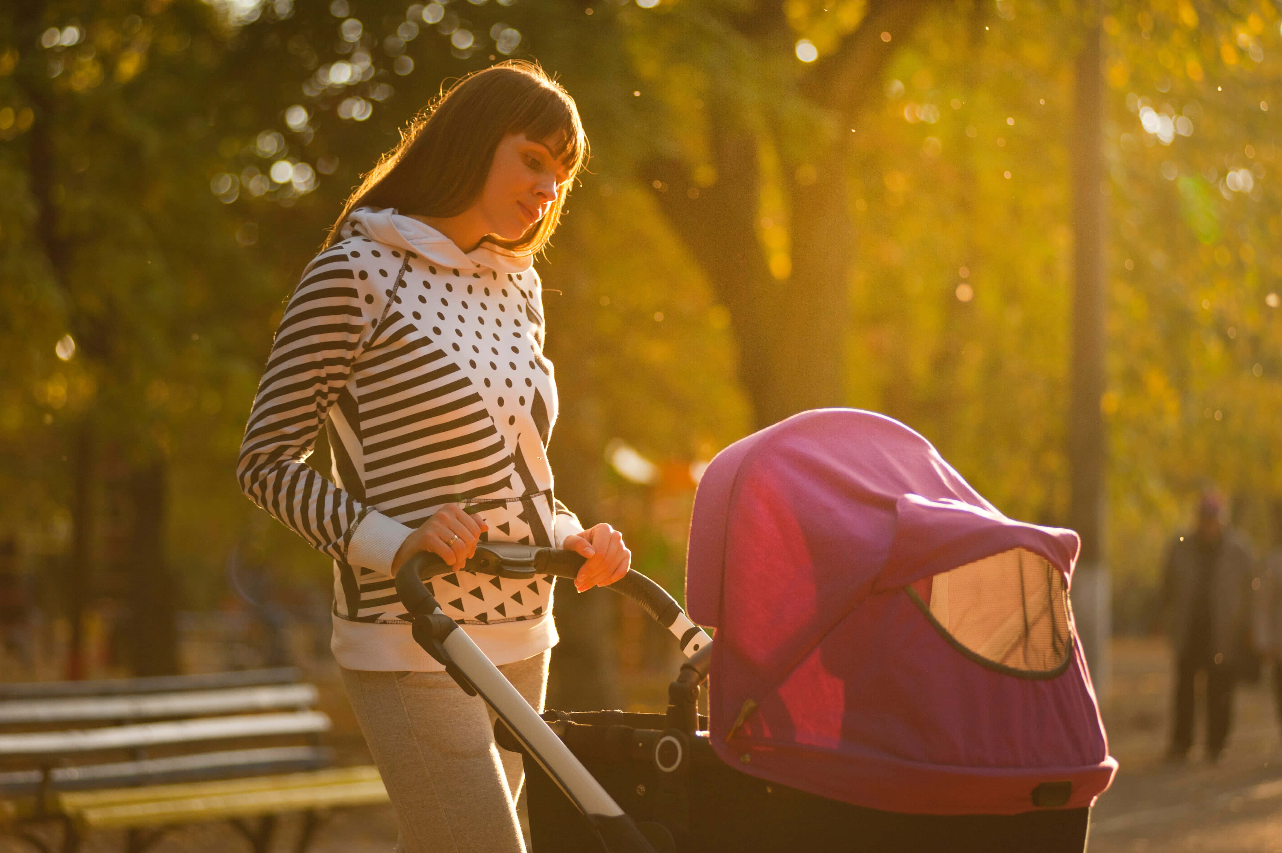 Mom 桌布 壁紙 — 一位女士推著紫色嬰兒車穿過陽光明媚的秋日公園，金色樹葉在她周圍旋轉。