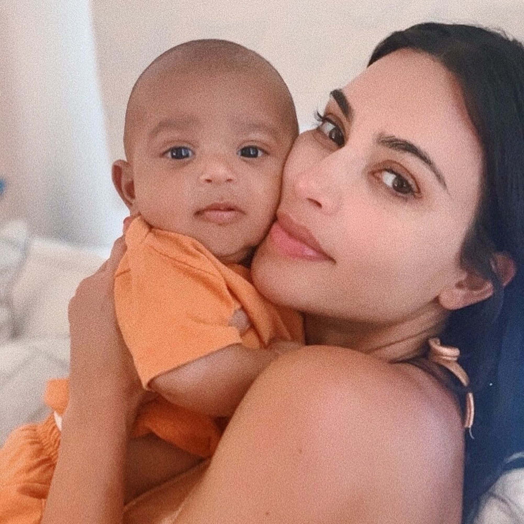 Baby 桌布 壁紙 — 一位女士溫柔地抱著一個穿著橙色連體衣的嬰兒，背景是柔和的白色。