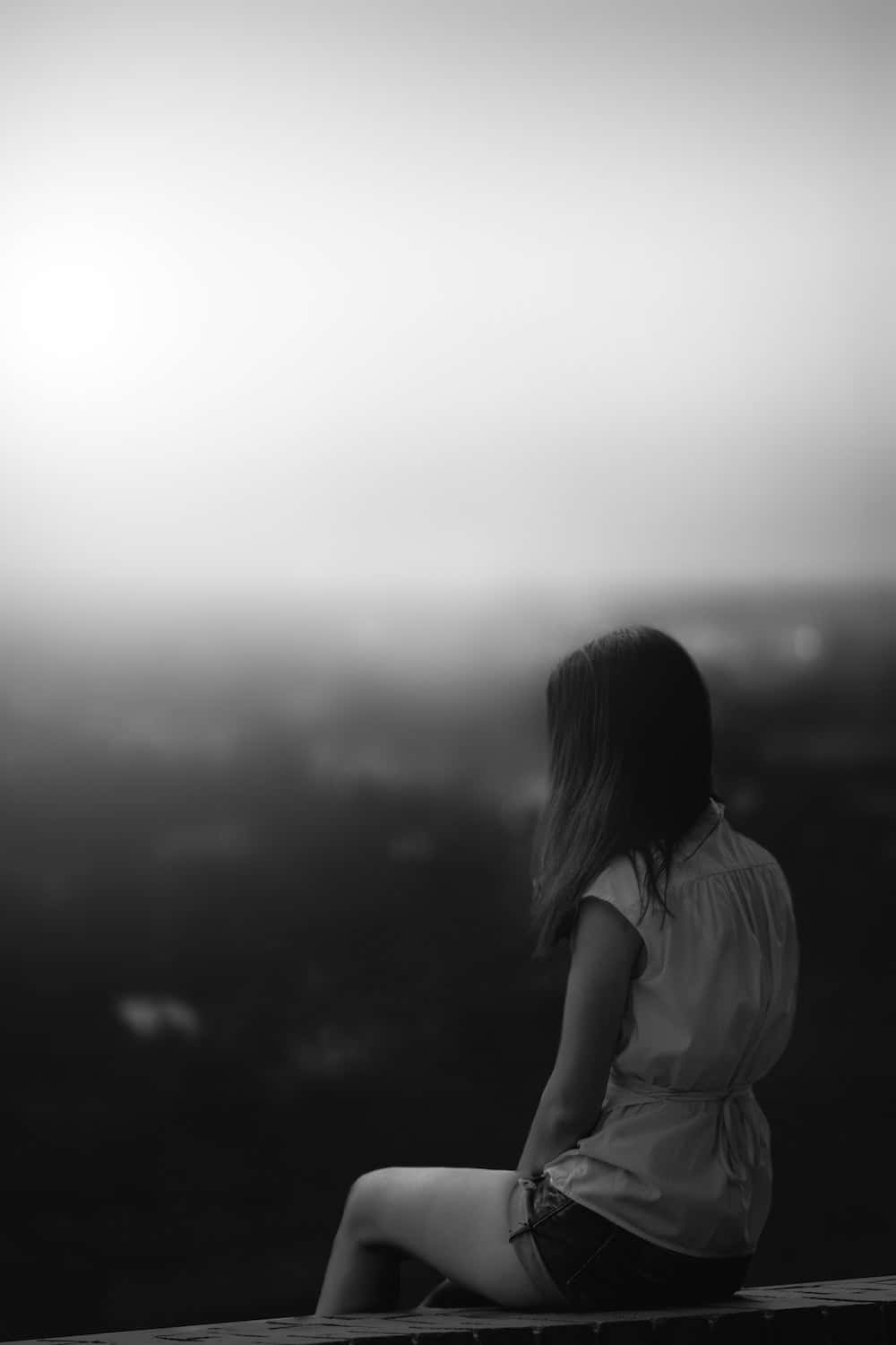 Sad Profile 桌布 壁紙 — 一位女士獨自坐在邊緣，俯瞰著朦朧的城市景觀，以單色呈現，她的剪影在柔和的光線中顯現。