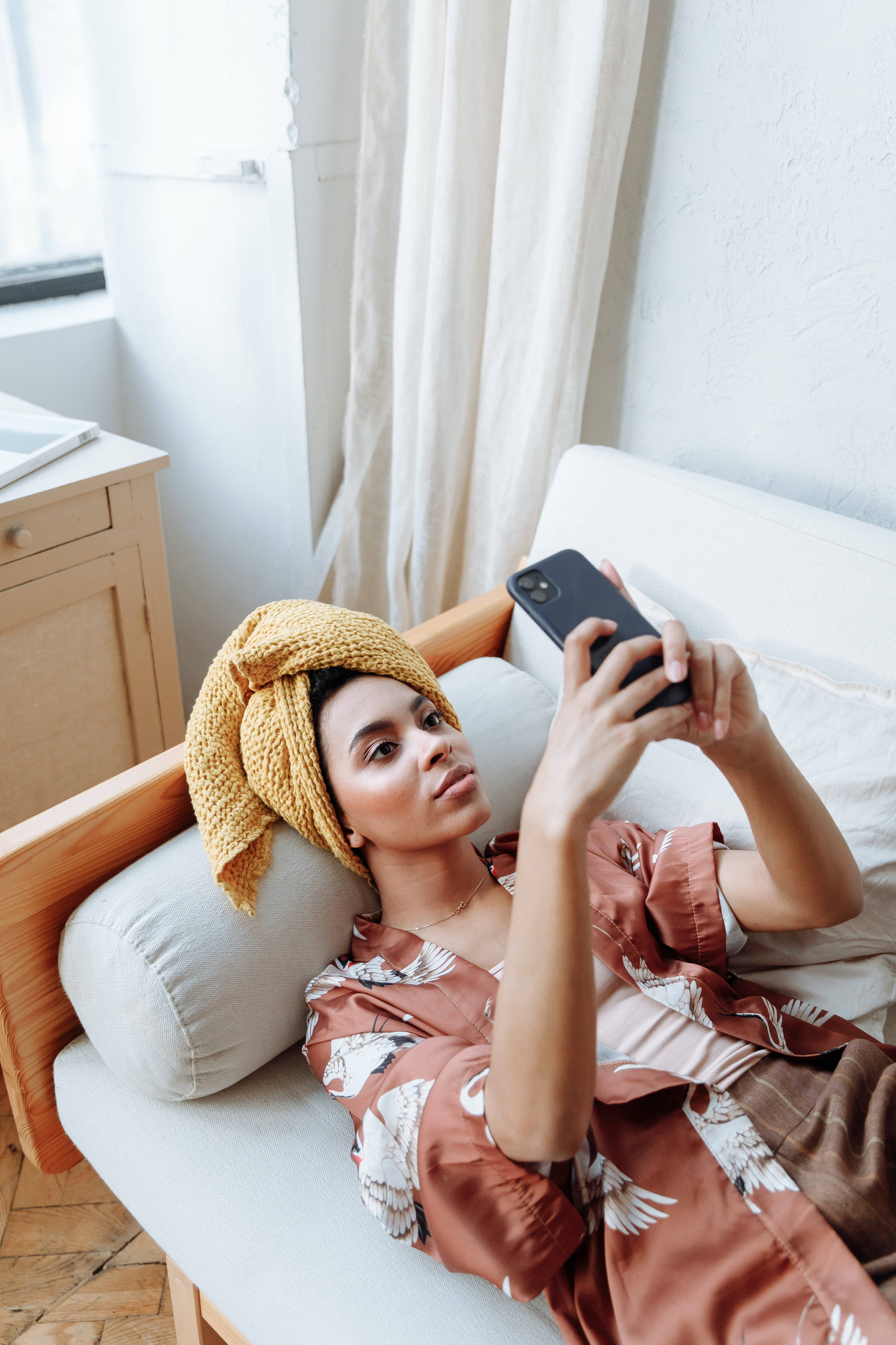 iPhone 桌布 壁紙 — 一位女士用黃色毛巾包裹頭部，在明亮的房間裏躺在白色沙發上專注地滑手機。