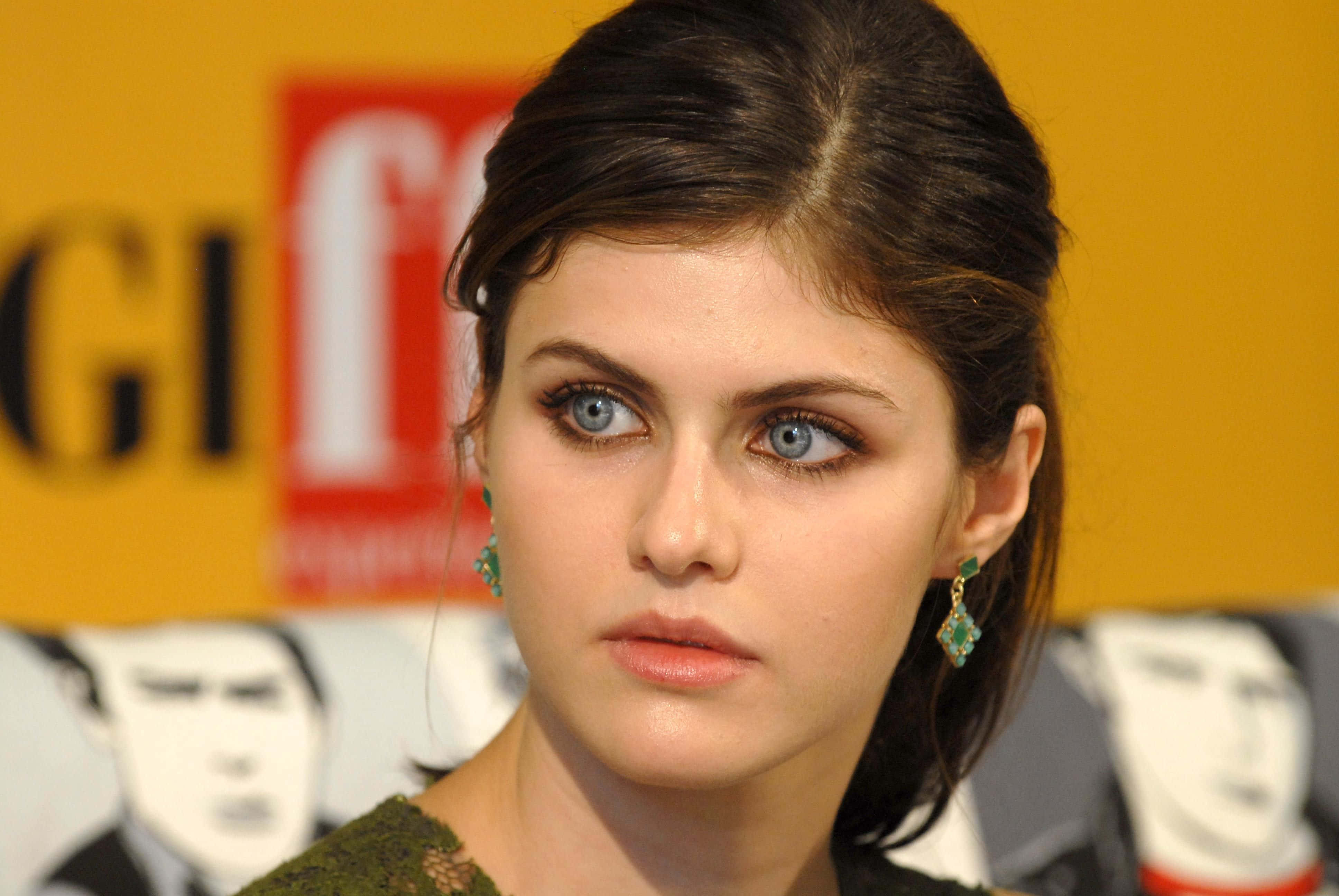 Alexandra Daddario 桌布 壁紙 — 一位女士的特寫肖像，擁有引人注目的藍色眼睛和向後梳理的深色頭髮，戴著綠色耳環，背景為黃色並帶有模糊的標誌。