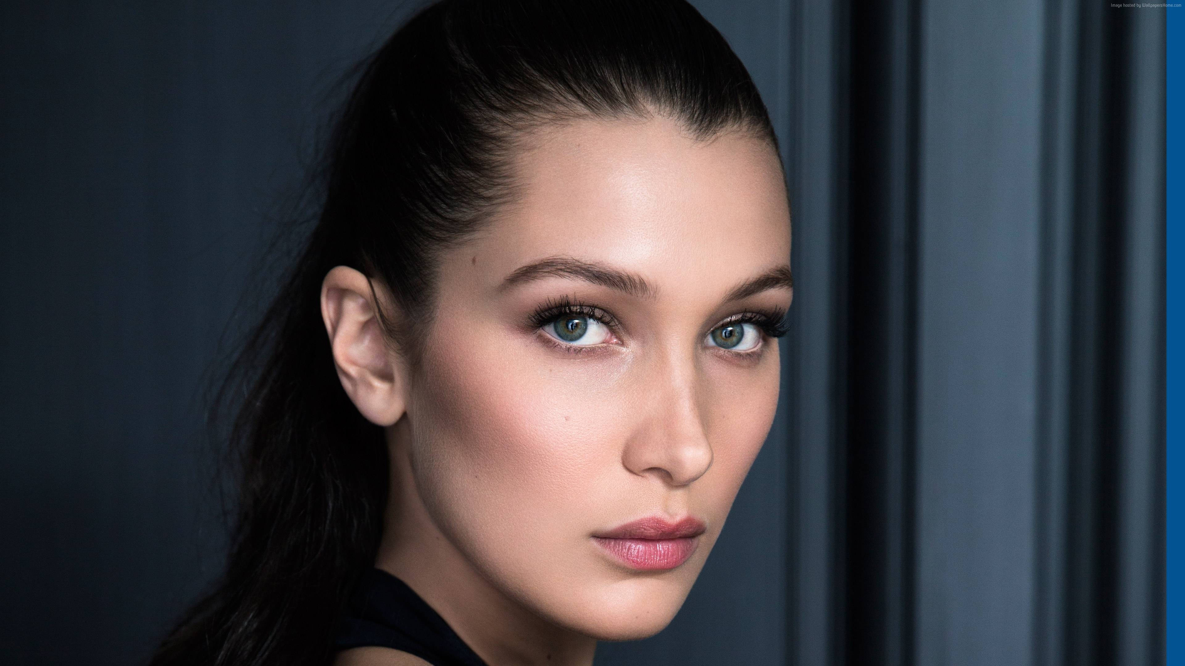 Bella Hadid 桌布 壁紙 — 一位女士的特寫肖像，擁有引人注目的藍色眼睛和深色頭髮向後梳，背景為柔和的灰色。