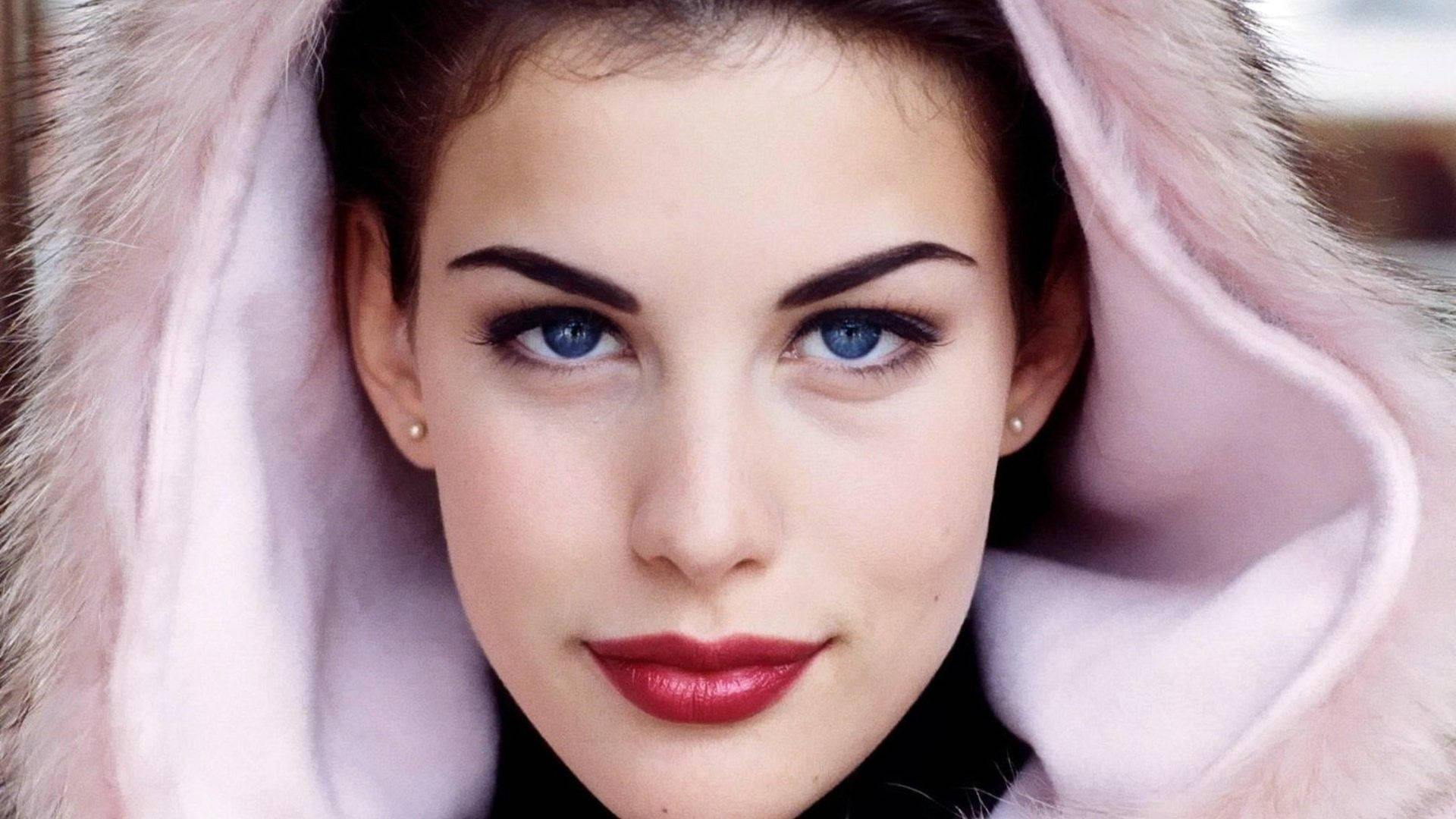 Liv Tyler 桌布 壁紙 — 一位女士的特寫肖像，擁有引人注目的藍色眼睛和鮮豔的紅唇，由柔軟的粉紅色毛絨帽兜框住。