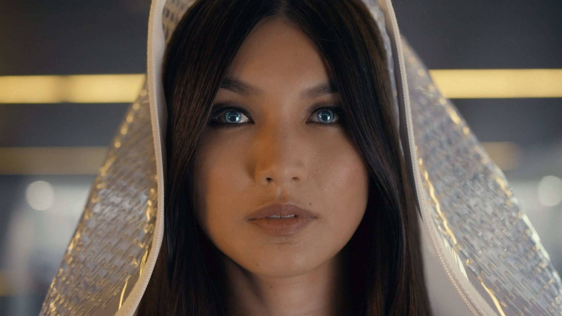 Gemma Chan 桌布 壁紙 — 一位女士的特寫肖像，擁有引人注目的藍色眼睛和深色頭髮，被閃閃發光的銀色連帽衣物所襯托。