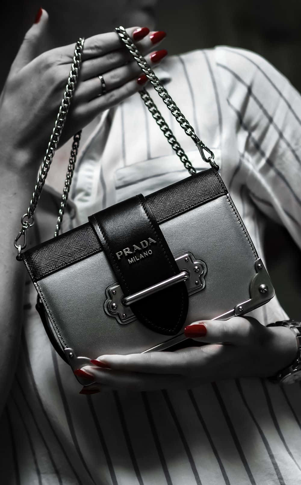 purse 桌布 壁紙 — 一位女士的雙手塗著紅色指甲，優雅地拿著一個帶鏈條的銀色Prada手袋，背景是單色條紋襯衫。