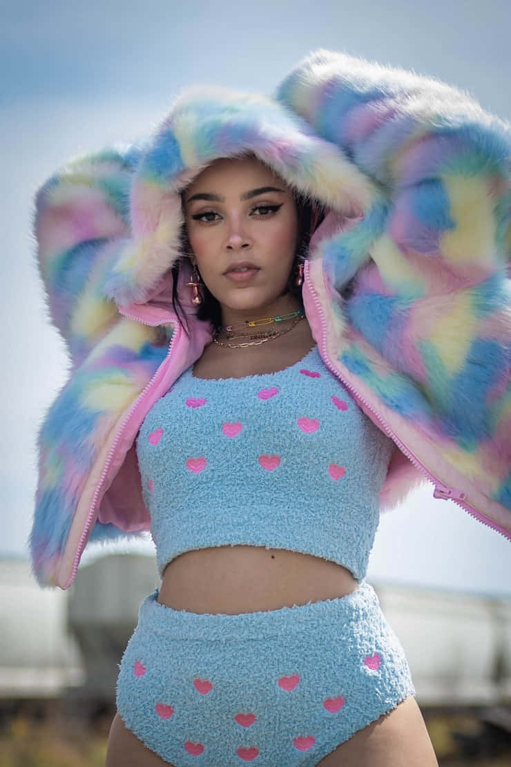 Doja Cat 桌布 壁紙 — 一位女士穿著充滿活力的彩虹毛絨連帽外套和淺藍色心形圖案服裝，背景是柔和天空。