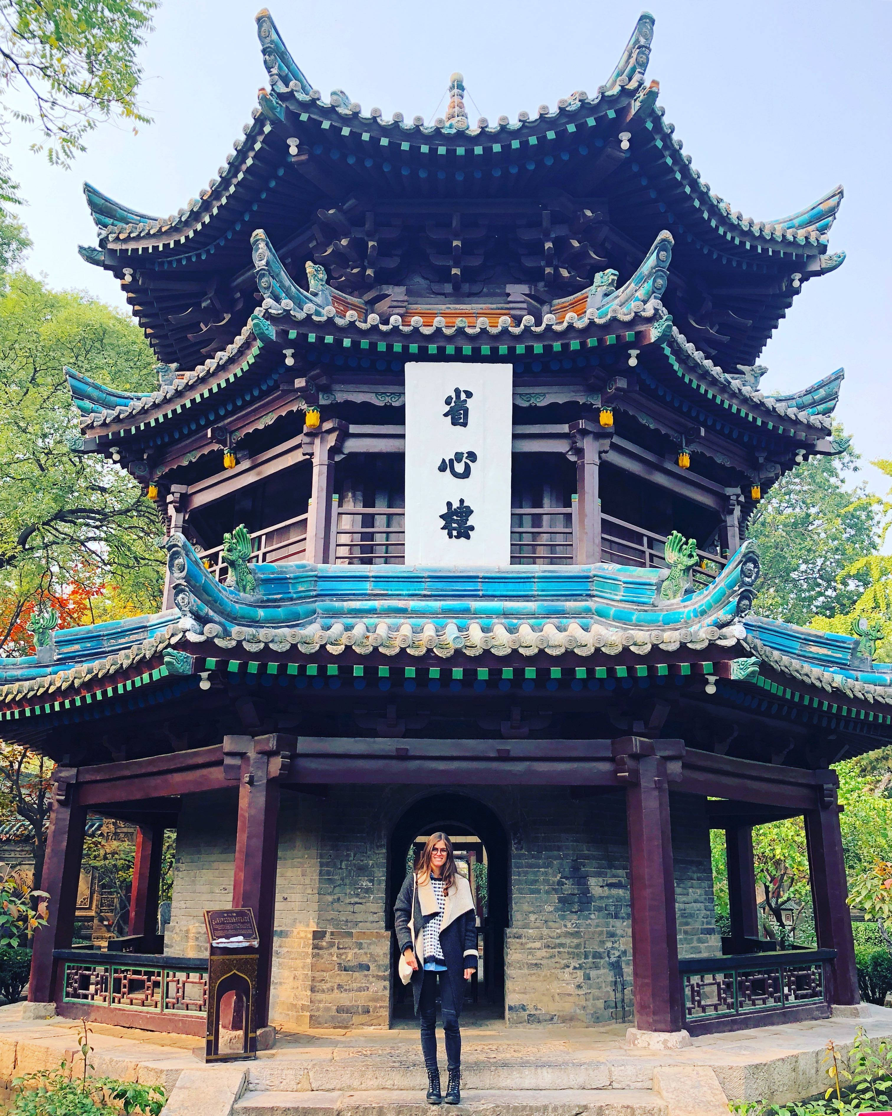 Xian 桌布 壁紙 — 一位女士站在傳統中式亭閣前，亭閣有彎曲的藍綠色瓦頂和木柱，周圍是茂密的綠樹，天空晴朗。