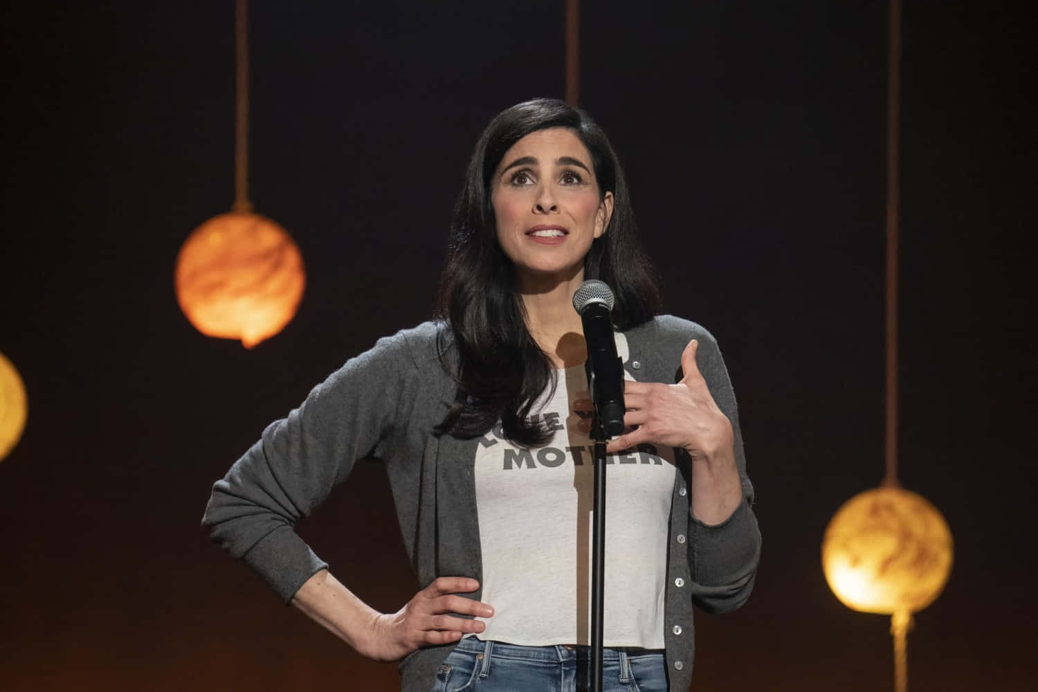 sarah silverman 桌布 壁紙 — 一位女士站在舞台上，手持麥克風，身後有溫暖的吊燈。