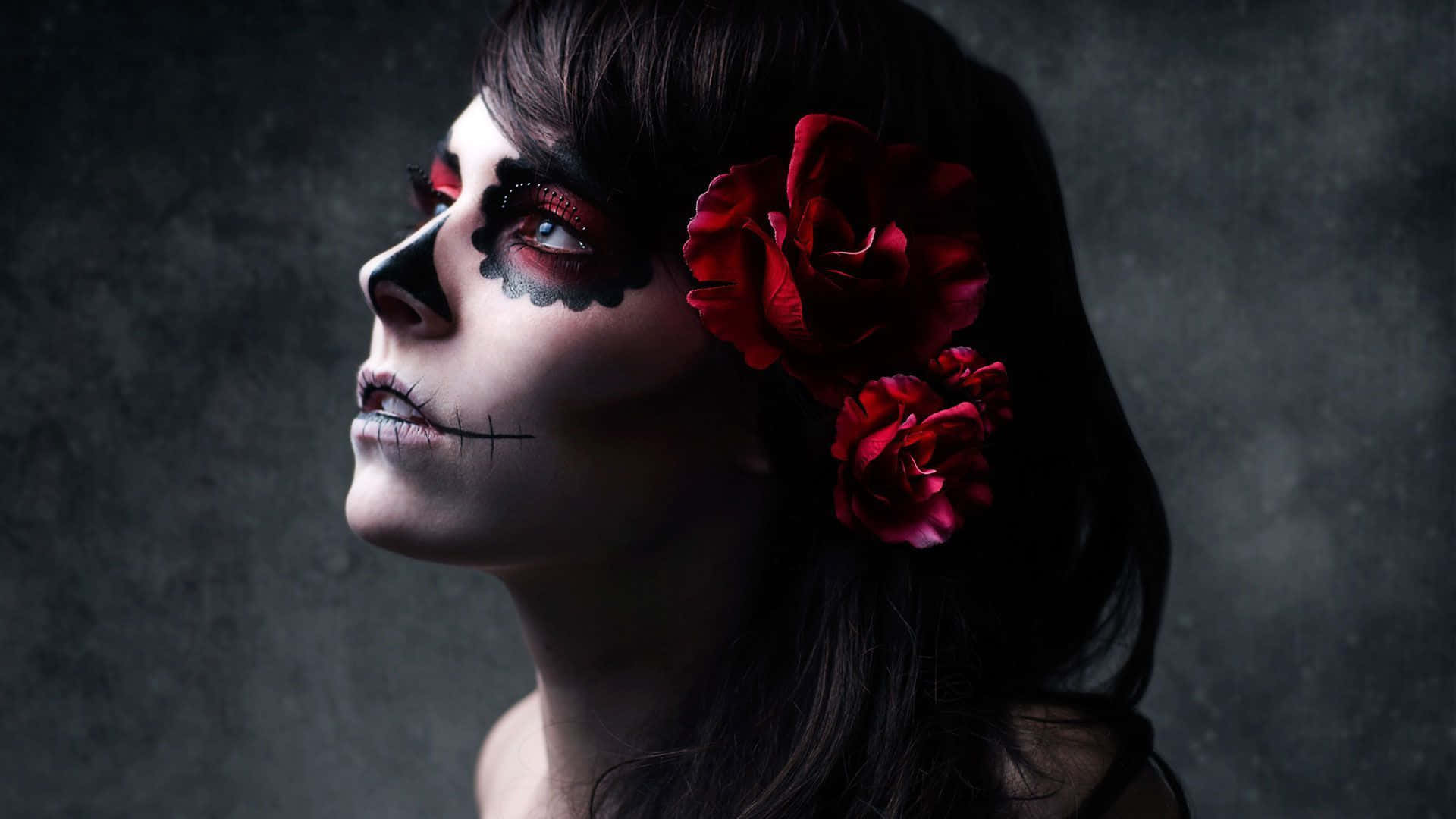 Day Of The Dead 桌布 壁紙 — 一位女士臉上畫著精緻的糖骷髏妝容，頭髮上插著紅玫瑰，在深色紋理背景前向上凝視。