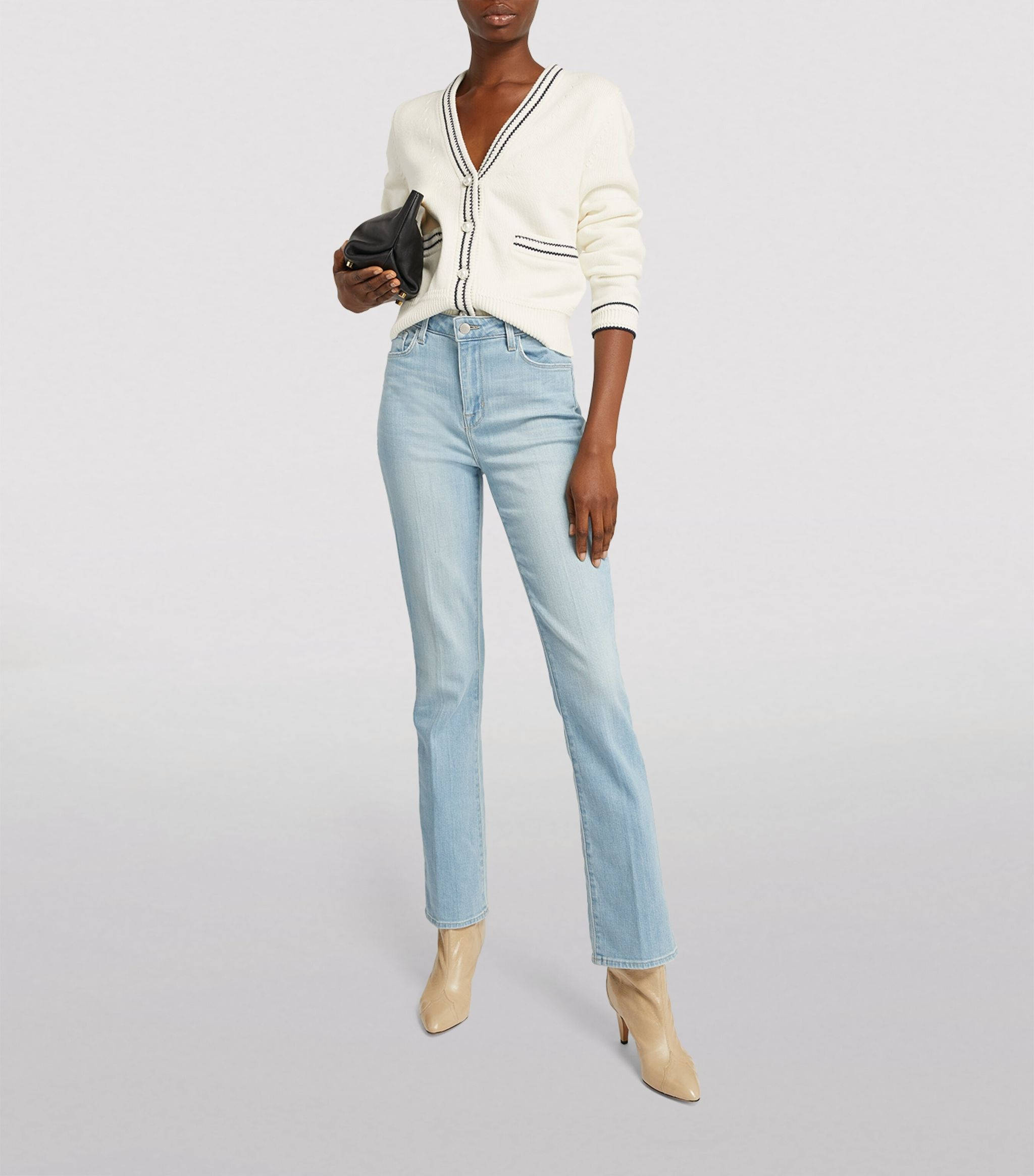 Denim 桌布 壁紙 — 一位女士自信地站立，身穿淺藍色牛仔褲、帶黑色飾邊的奶油色開襟衫和米色踝靴，手持黑色手拿包，背景為純白色。