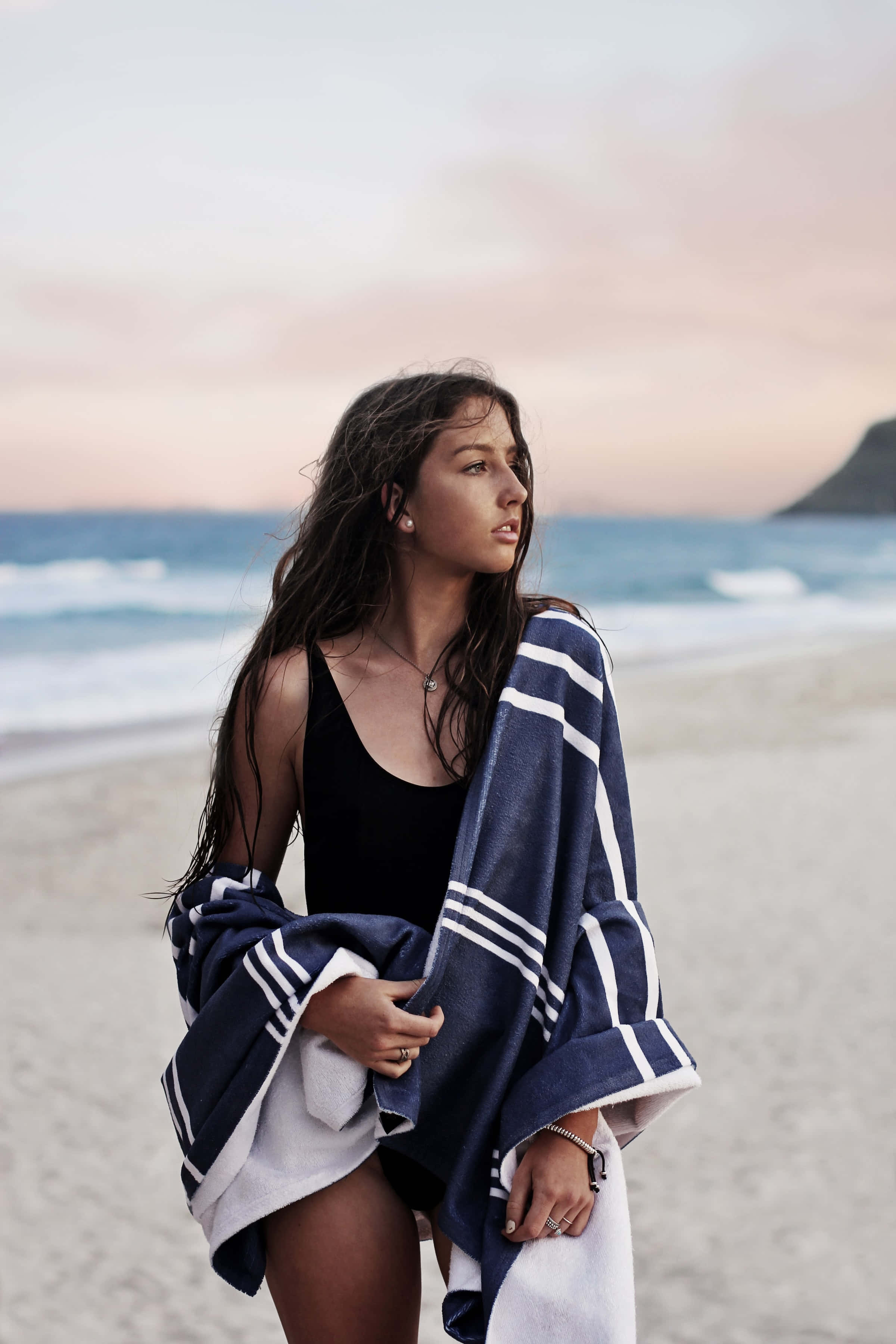 Swimsuit 桌布 壁紙 — 一位女士裹著海軍藍白條紋毛巾，站在日落時分的沙灘上，身後是海浪。