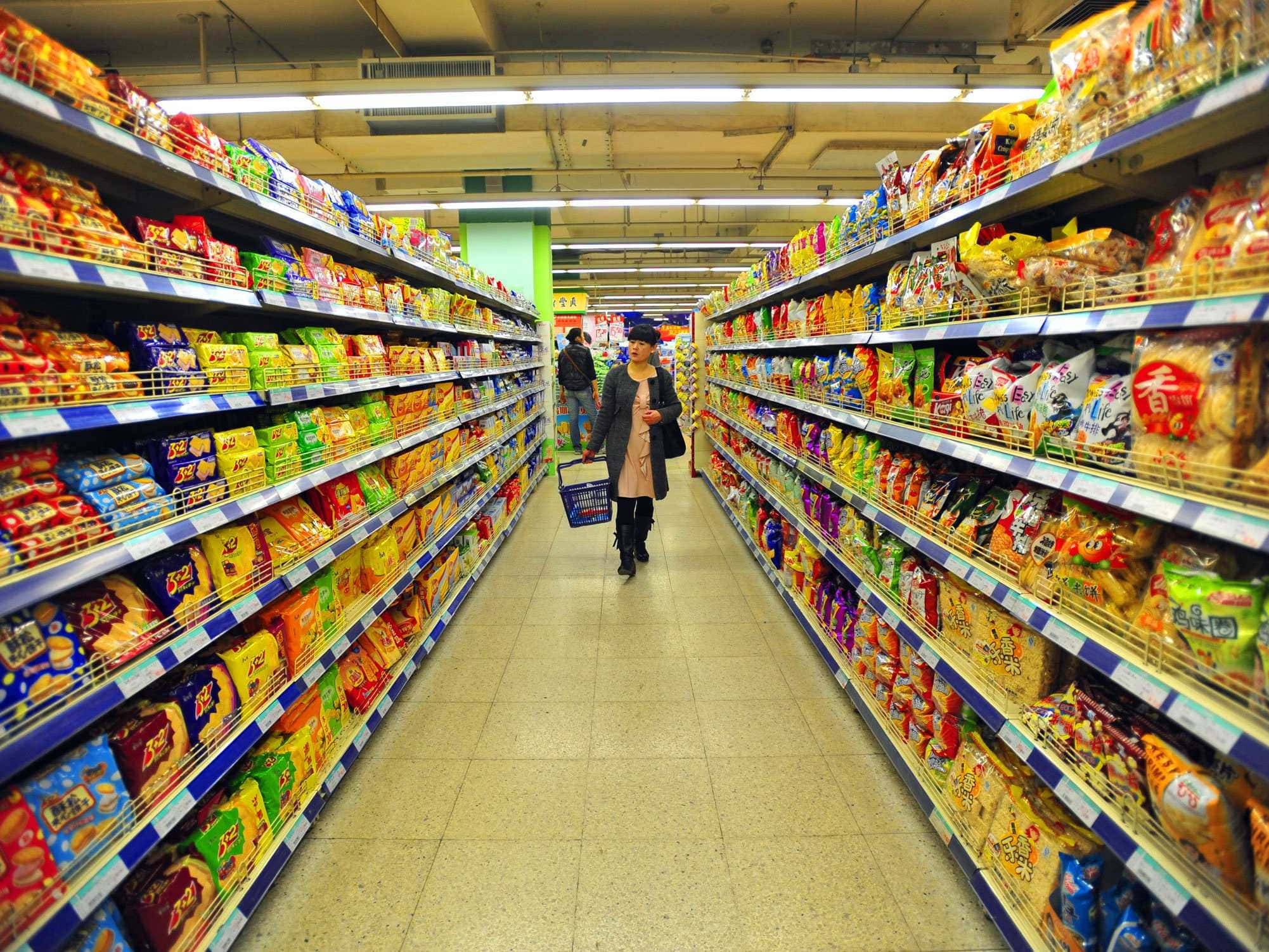 supermarket 桌布 壁紙 — 一位女士走在光線明亮的超市通道上，兩旁擺滿了色彩繽紛的零食包裝。
