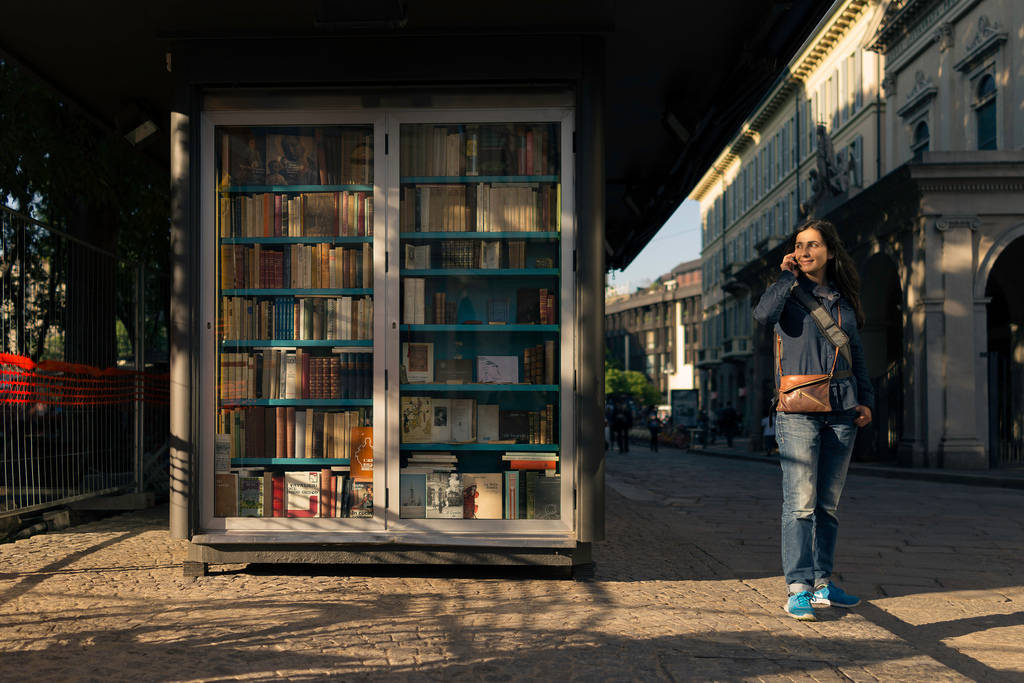 Woman 桌布 壁紙 — 一位女士走過一個玻璃面的書報攤，裡面擺滿了書籍，位於陽光普照的鵝卵石街道上。