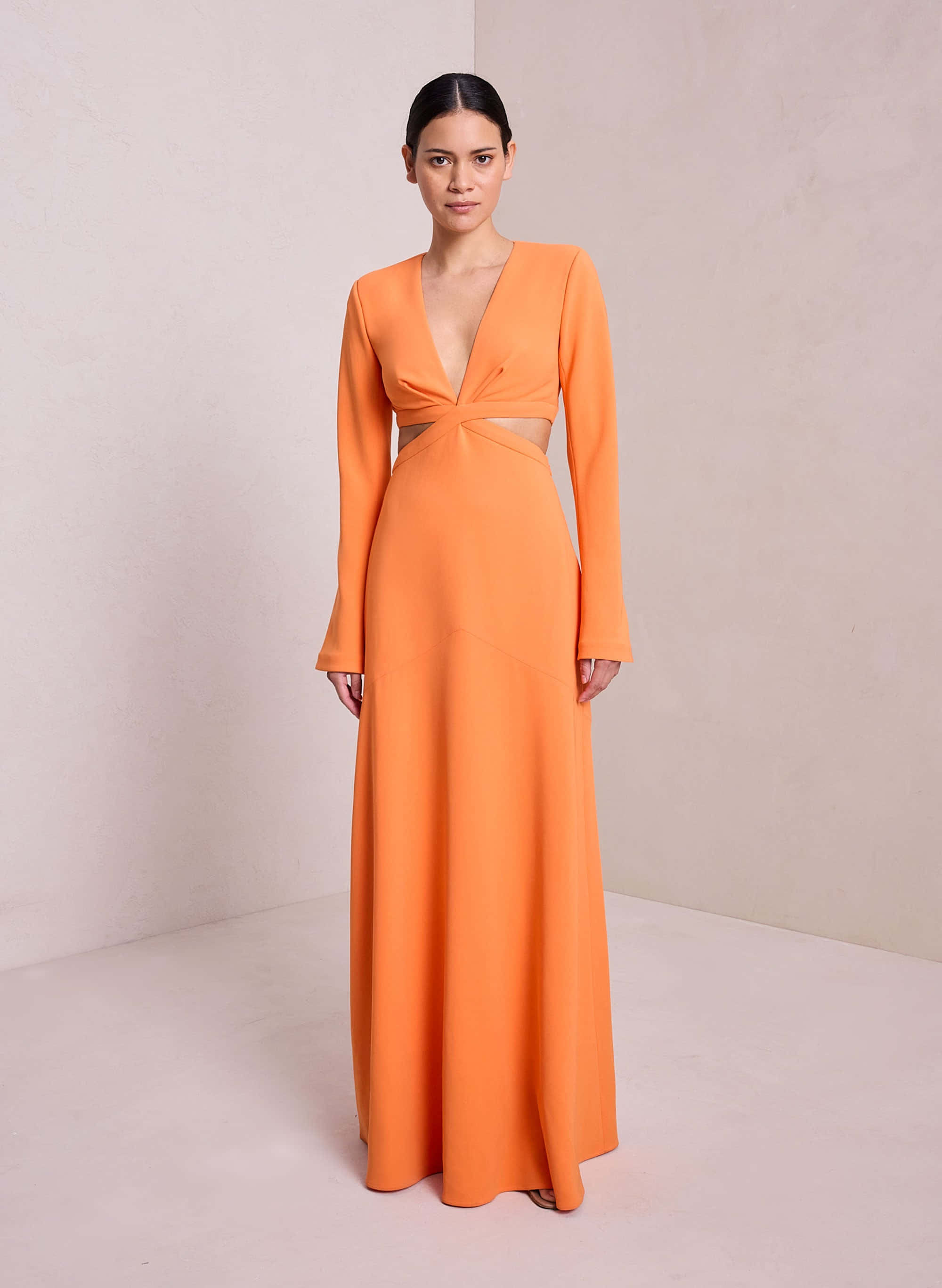 Long Dress 桌布 壁紙 — 一位女士身穿鮮豔的橙色落地長裙，腰部扭結設計，長袖，在簡約的米色工作室背景前優雅站立。