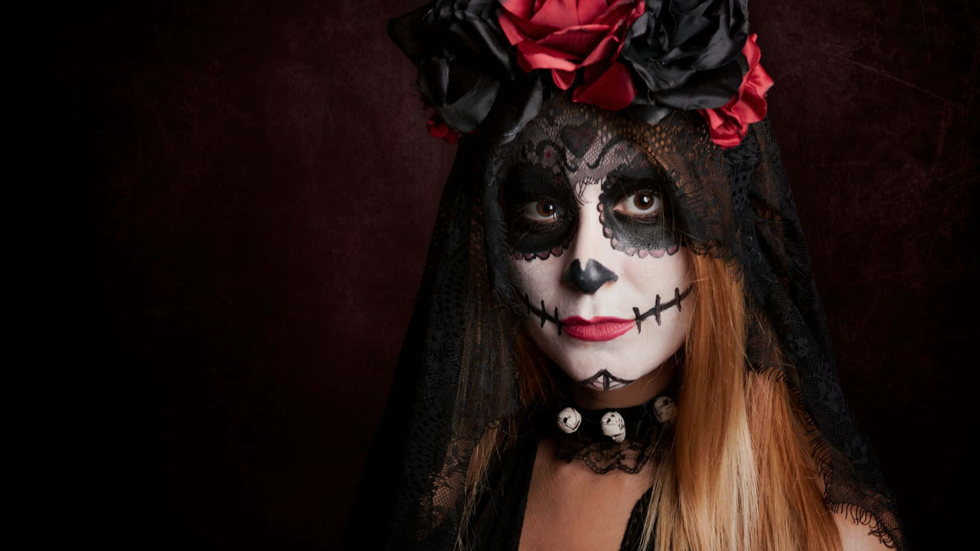 Halloween Costumes 桌布 壁紙 — 一位女性，臉上畫著精緻的糖骷髏妝容，戴著飾有紅黑玫瑰的黑色蕾絲面紗，在深酒紅色背景下直視鏡頭。