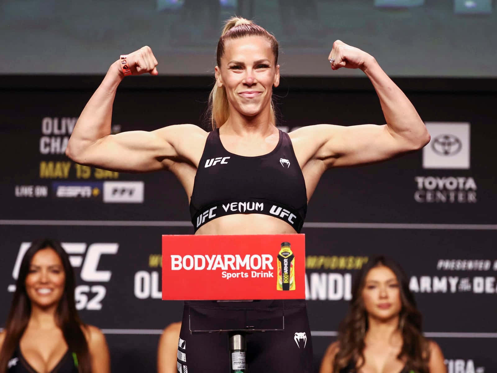 桌布 壁紙 — 一位女性UFC拳手在稱重活動中自信地微笑，彎曲她肌肉發達的手臂。