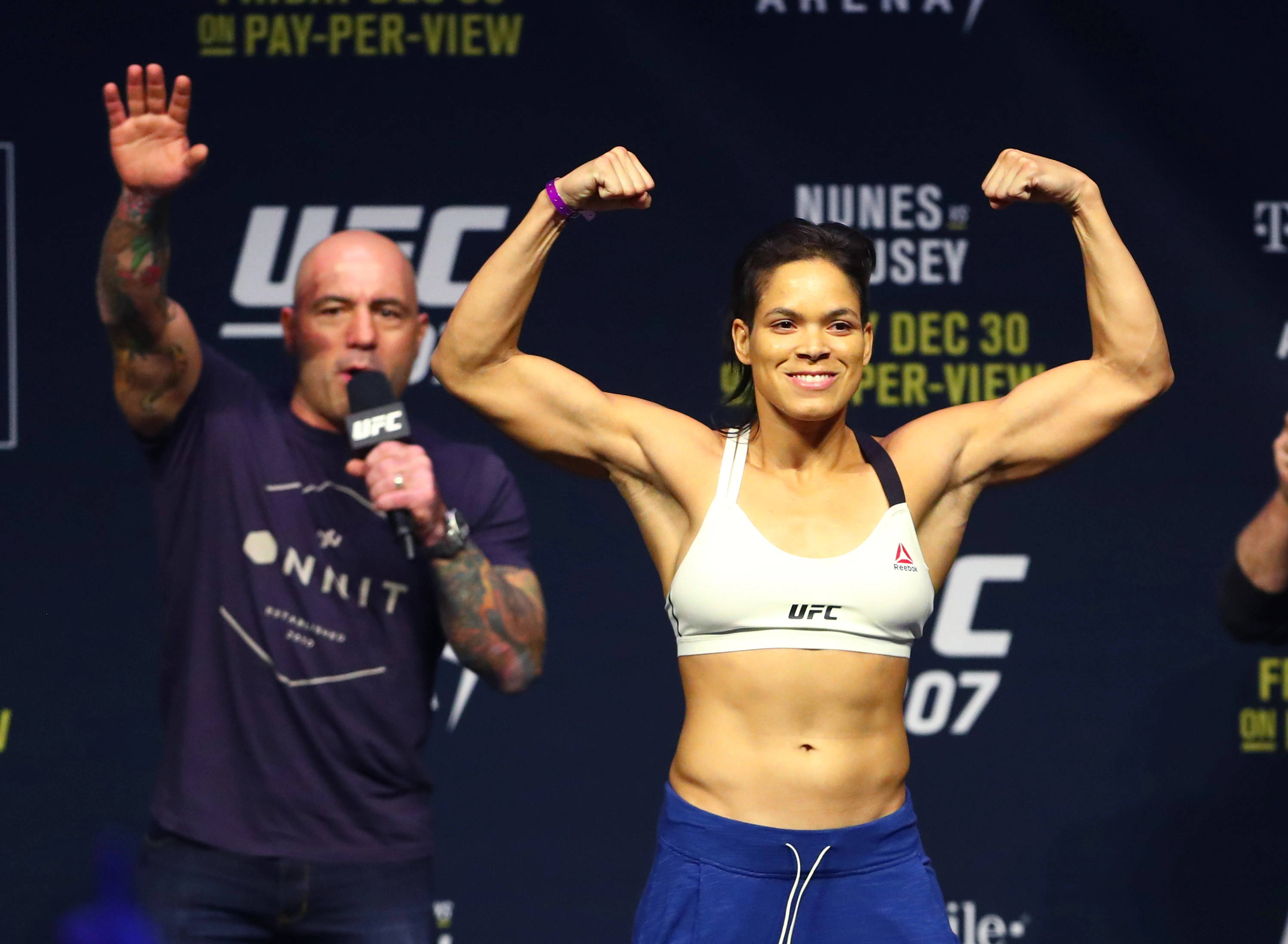 MMA 桌布 壁紙 — 一位女性UFC拳手在稱重活動中自信地微笑，彎曲她肌肉發達的手臂，而一位有刺青的男子在她旁邊拿著麥克風。