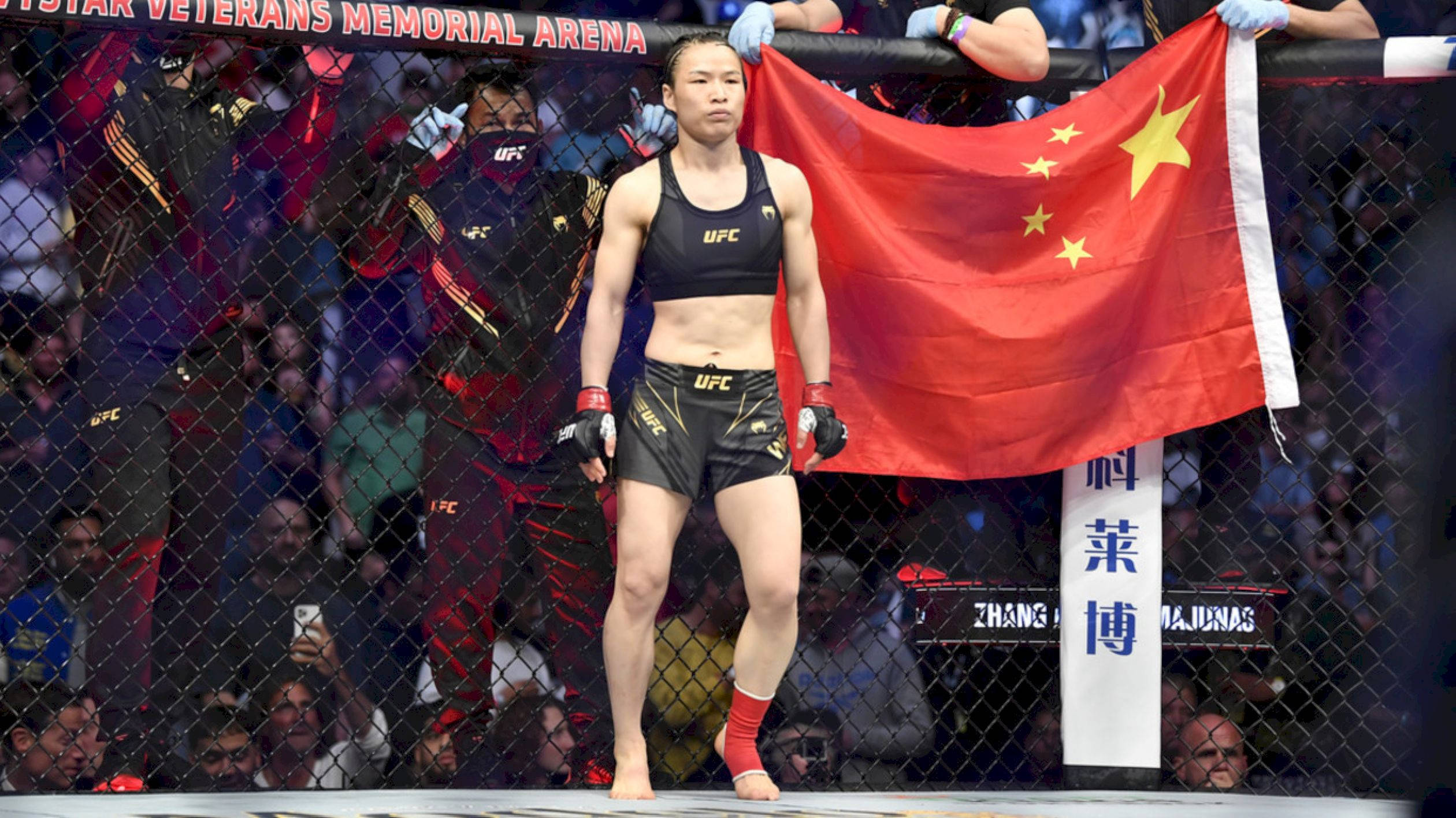 Athletic 桌布 壁紙 — 一位女性UFC拳手自信地站在八角籠中，手持中國國旗，粉絲們在圍欄外觀看。