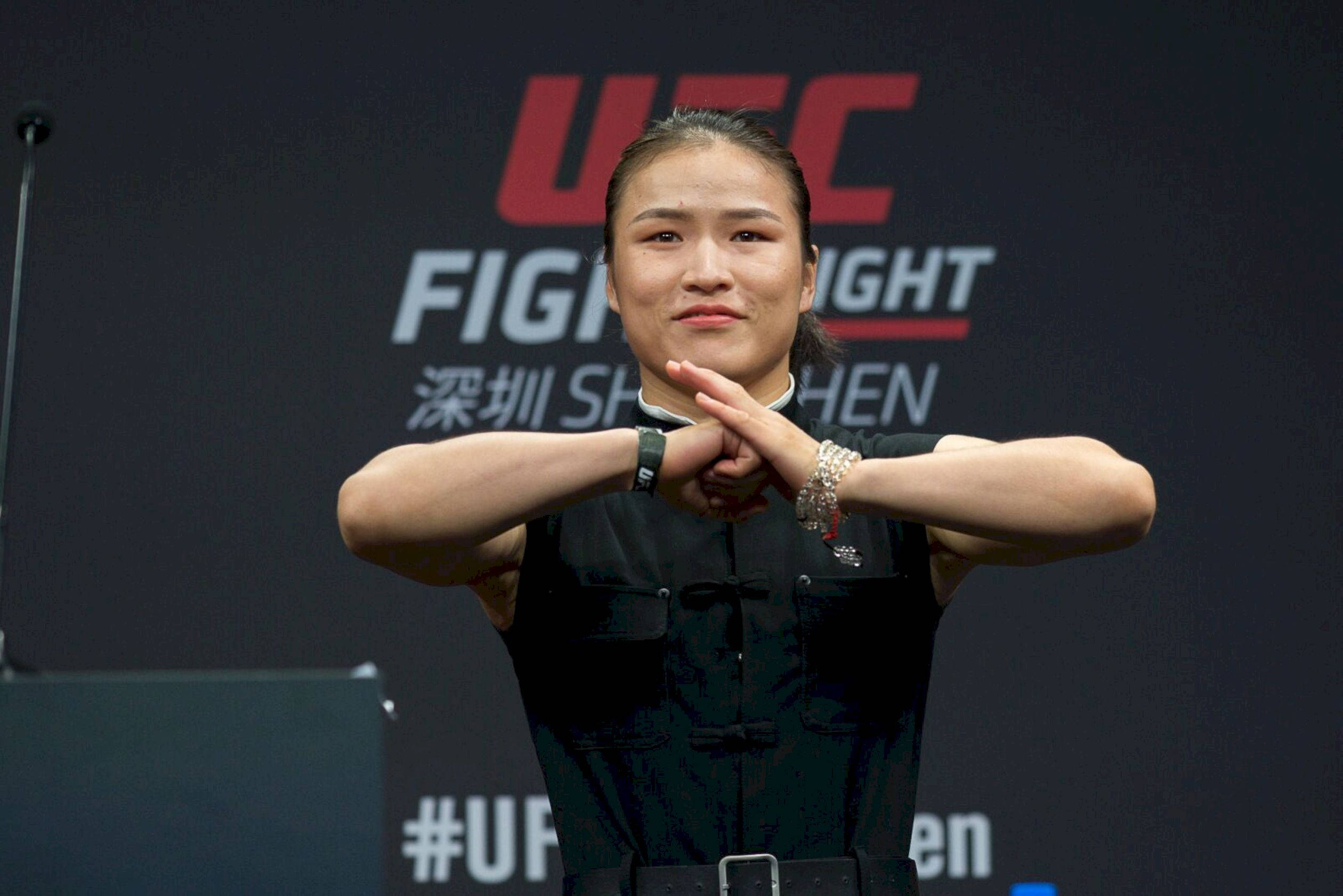 Athletic 桌布 壁紙 — 一位女性UFC拳手自信地站在記者會上，雙臂交叉在身前，身穿黑色服裝。
