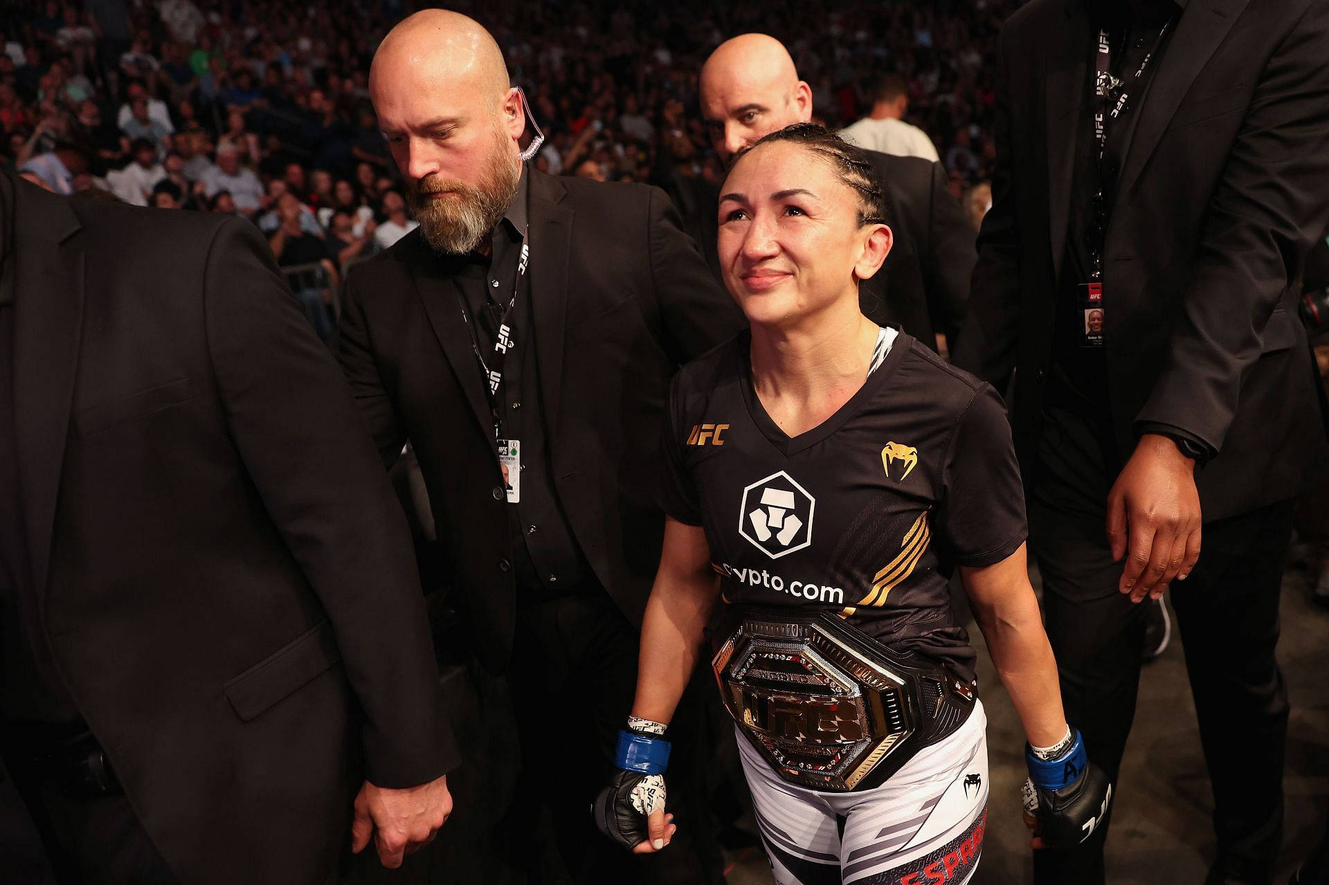 MMA 桌布 壁紙 — 一位女性UFC拳手自豪地微笑著，戴著冠軍腰帶穿過擁擠的競技場。