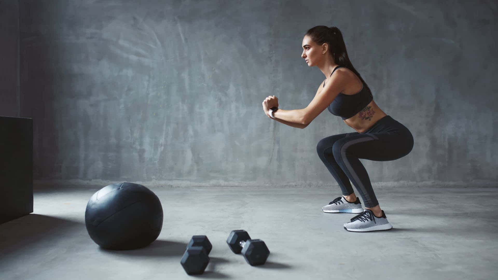 Dumbbell 桌布 壁紙 — 一位女性在極簡風格的健身房中，使用啞鈴和健身球在混凝土地板上對著灰色牆壁進行深蹲。