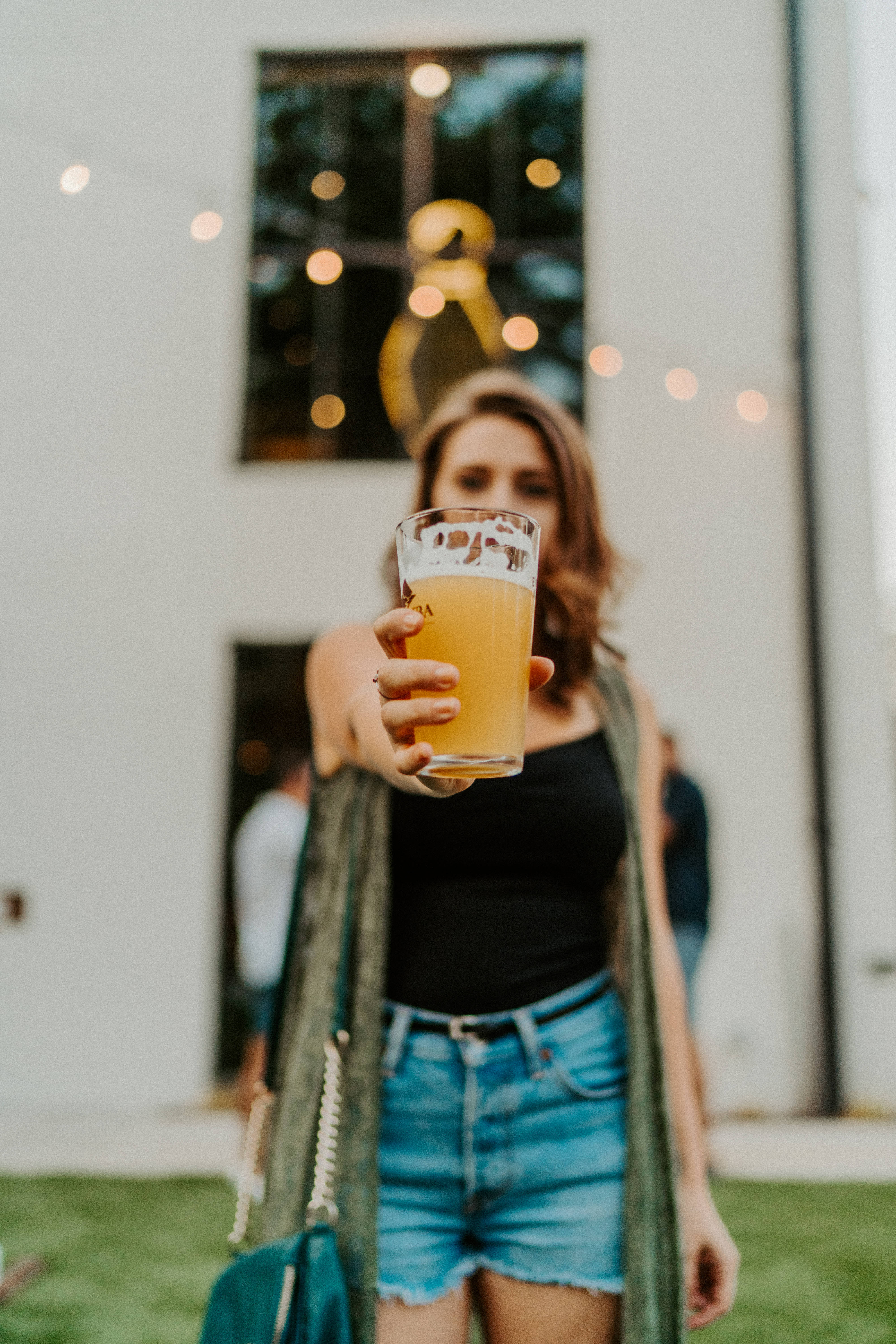 Beer 桌布 壁紙 — 一位女性在陽光普照的戶外庭院中，身後有燈串和現代建築，向鏡頭遞出一杯泡沫啤酒。