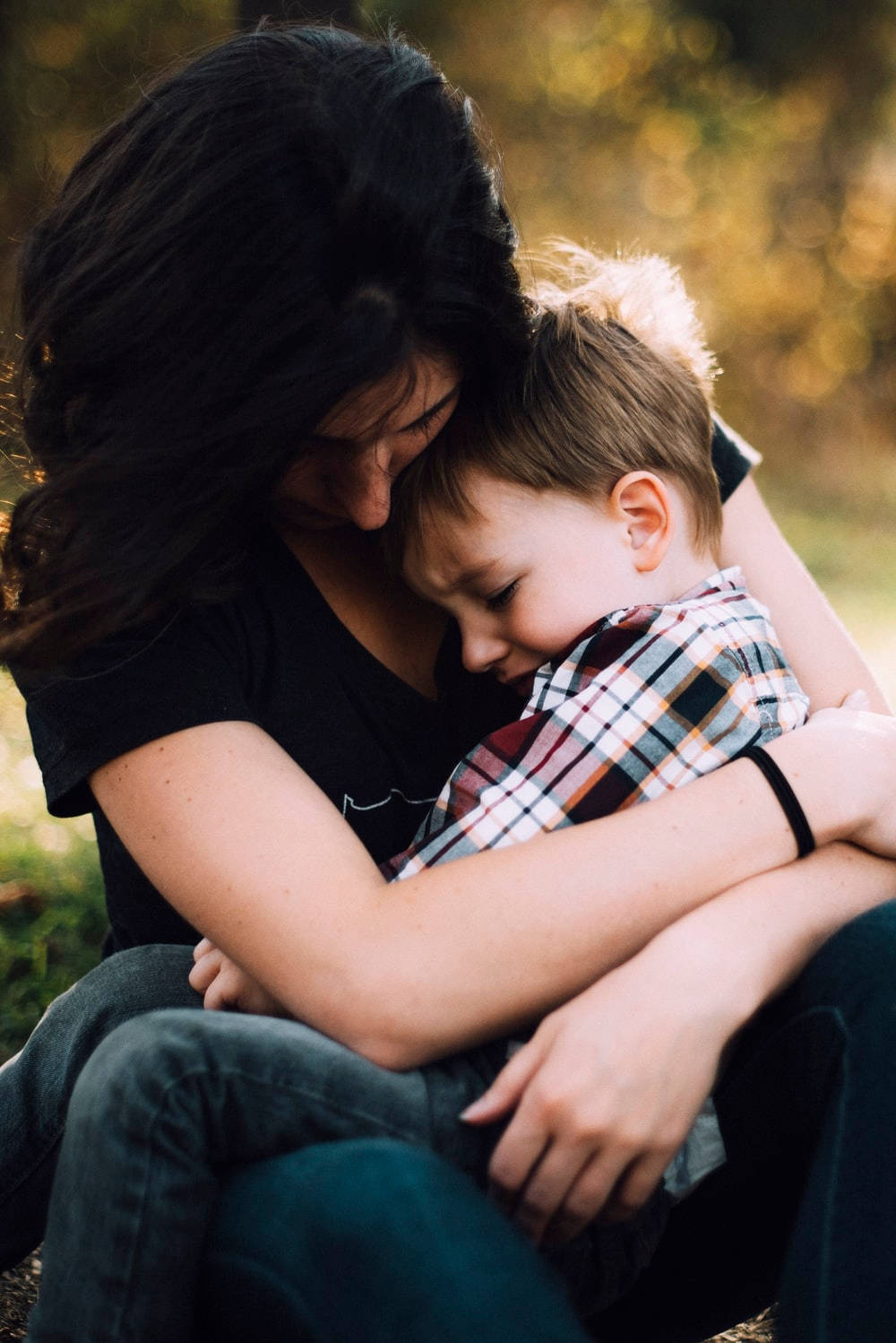 Family 桌布 壁紙 — 一位女性在陽光普照的戶外環境中，溫柔地擁抱著一個熟睡的孩子，背景是金黃色的樹葉。