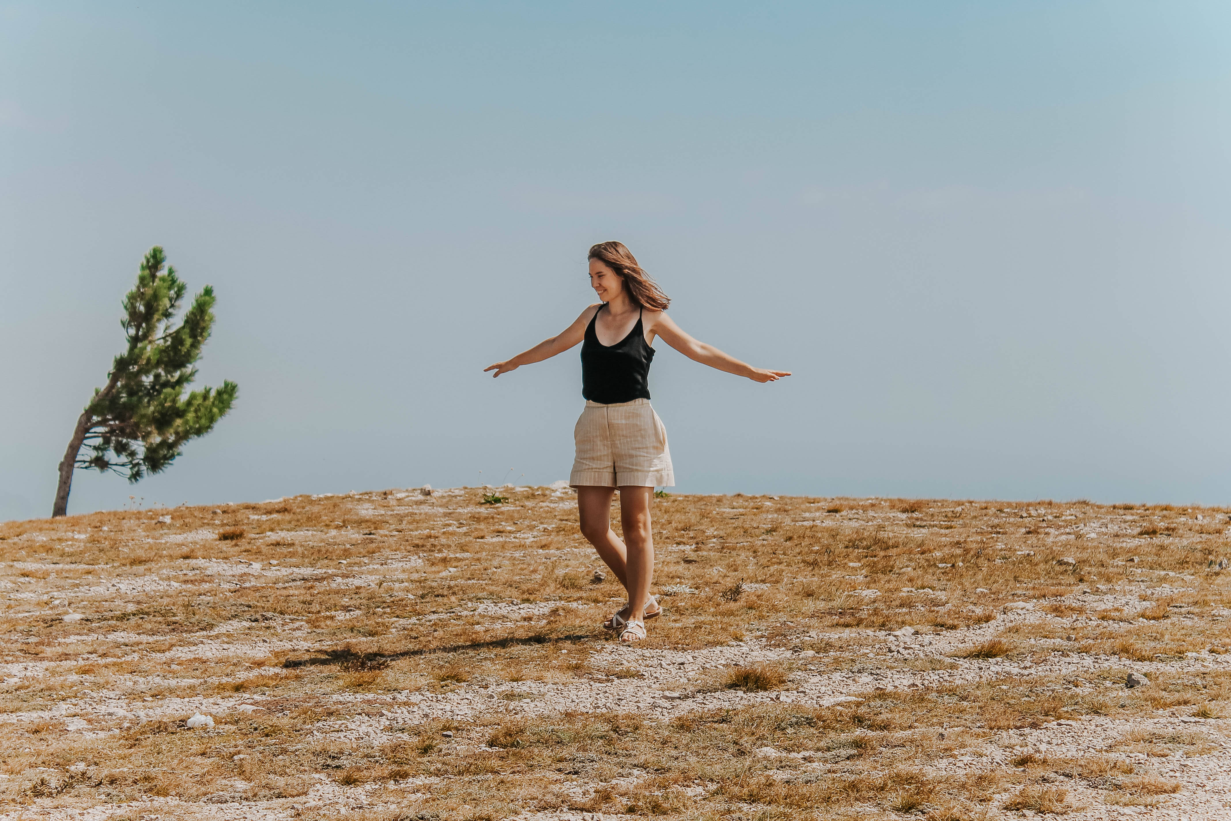 Forever Alone 桌布 壁紙 — 一位女性張開雙臂，愉快地走過陽光明媚、乾燥的草地山坡，天空湛藍，附近有一棵孤獨的松樹。
