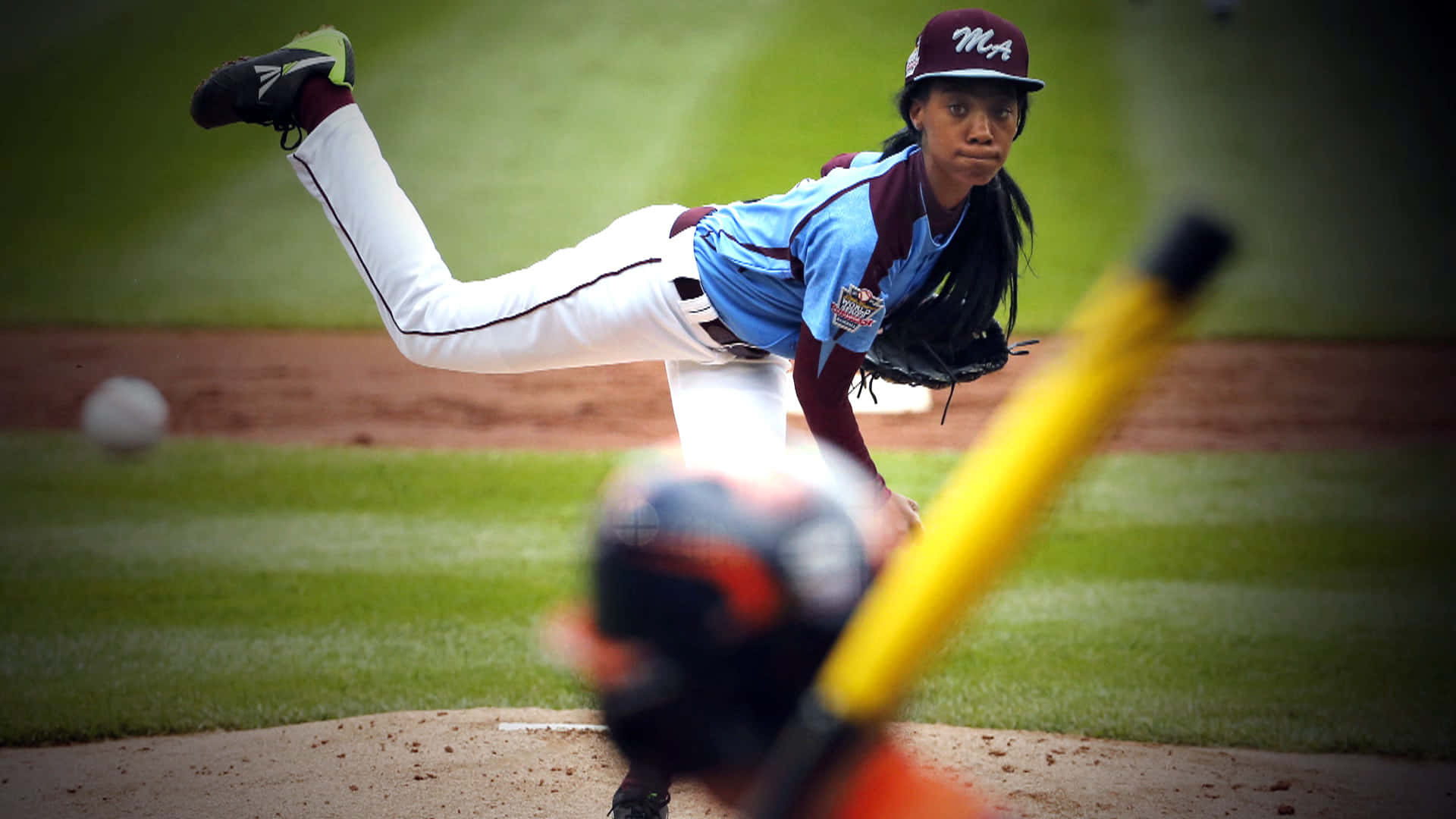 Little League Baseball 桌布 壁紙 — 一位女性棒球投手在陽光明媚的球場上投球，前景有模糊的打者和球棒。