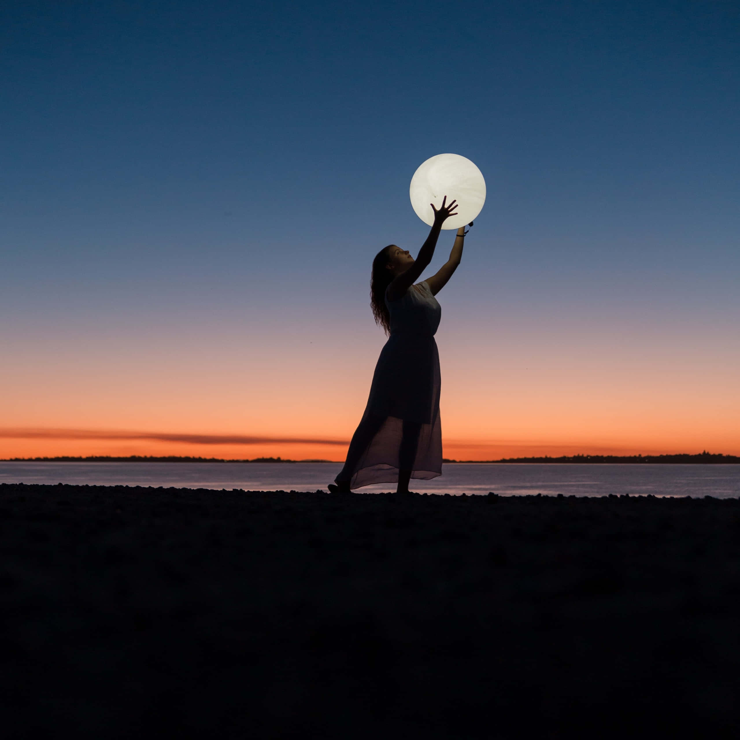 Beach Photography 桌布 壁紙 — 一位女性的剪影在暮色中的沙灘上向上伸展，手持發光球體，映襯著漸變的天空。