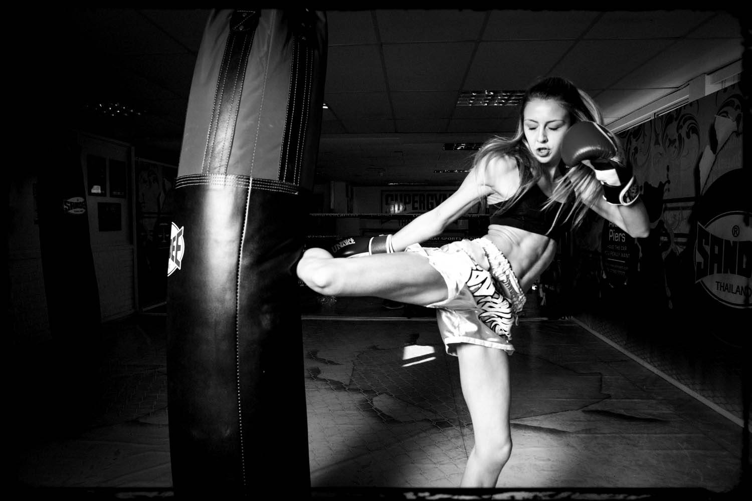 Athletic 桌布 壁紙 — 一位女拳擊手在昏暗的健身房中對沙包使出高踢，以戲劇性的黑白畫面捕捉。