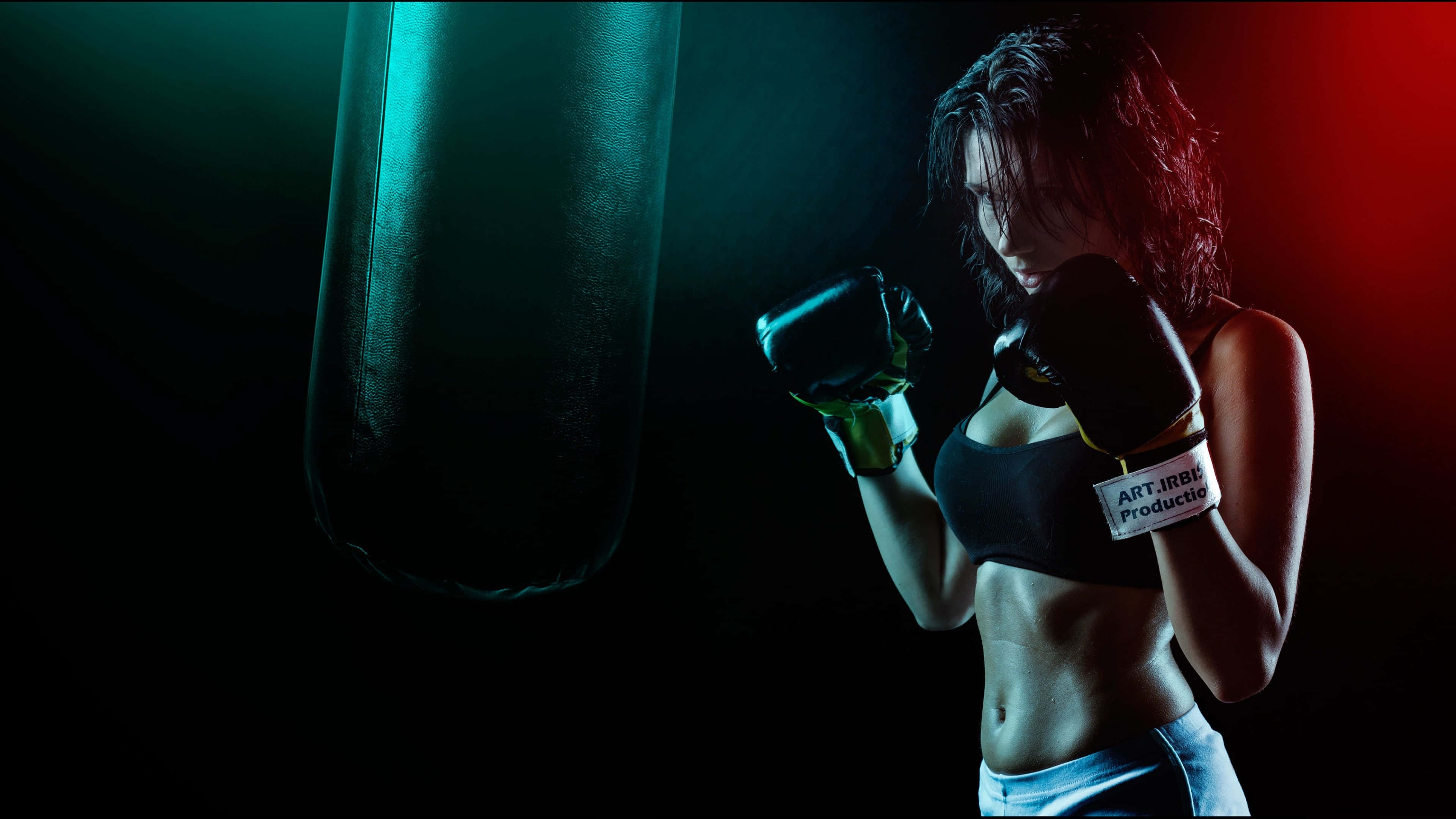 Boxing 桌布 壁紙 — 一位女拳擊手戴著黑色手套和運動內衣，在戲劇性的紅色和藍綠色燈光下，準備好站在重型沙包旁。