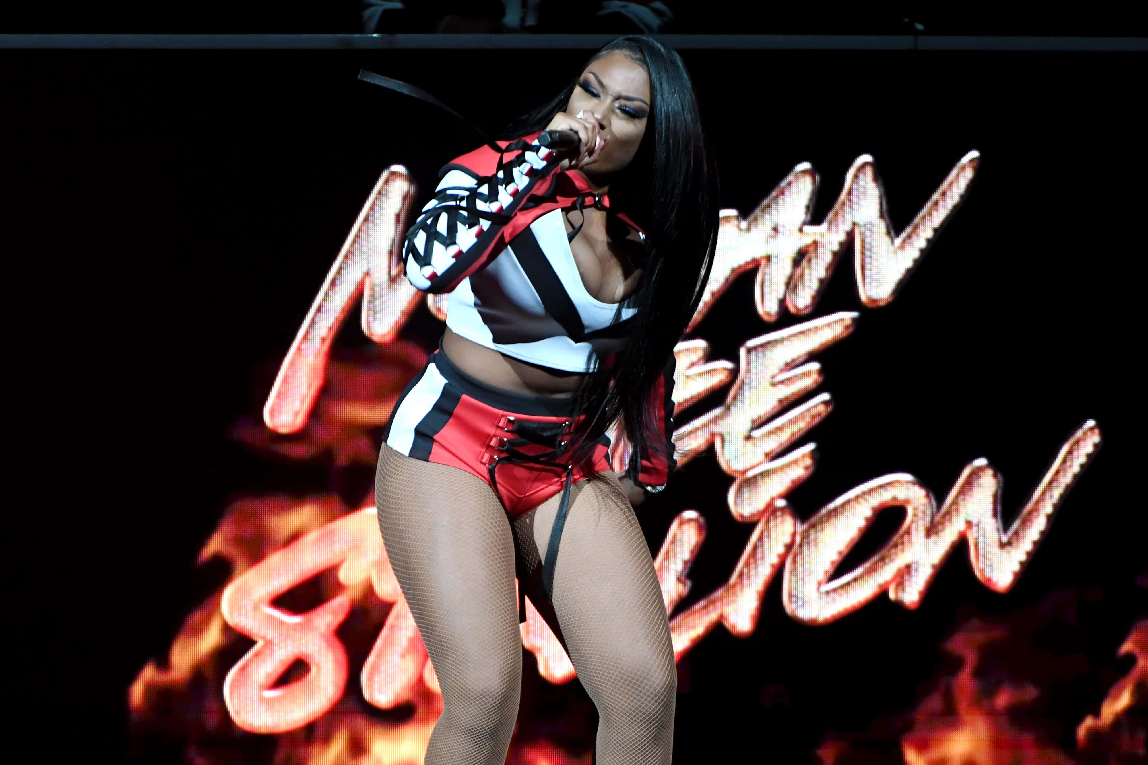 Megan Thee Stallion 桌布 壁紙 — 一位女歌手穿著紅、白、黑三色服裝，在熾熱的背景燈光下於舞台上表演。
