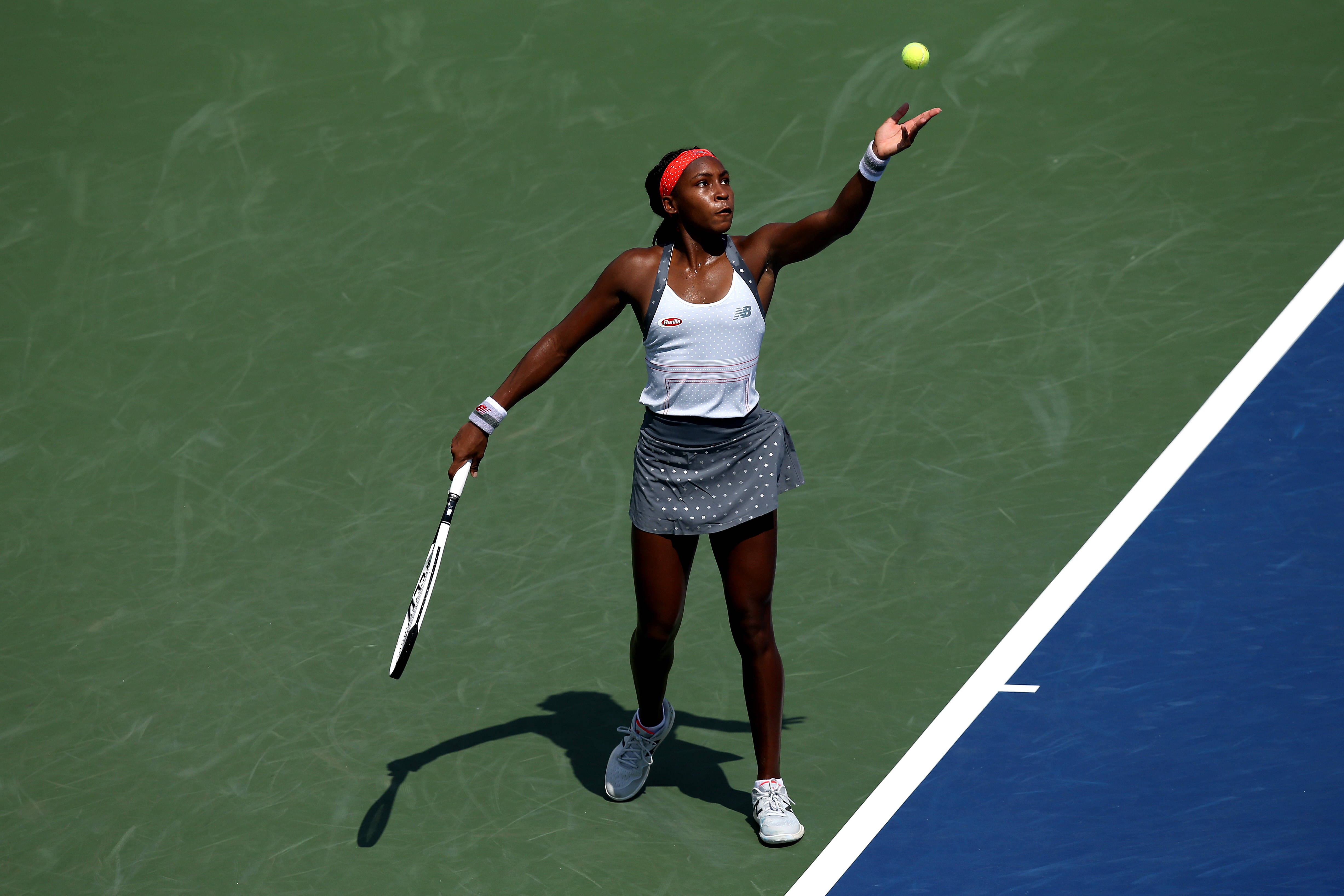 Coco Gauff 桌布 壁紙 — 一位女網球運動員在綠色和藍色的球場上，在明亮的陽光下將黃色球拋向空中。