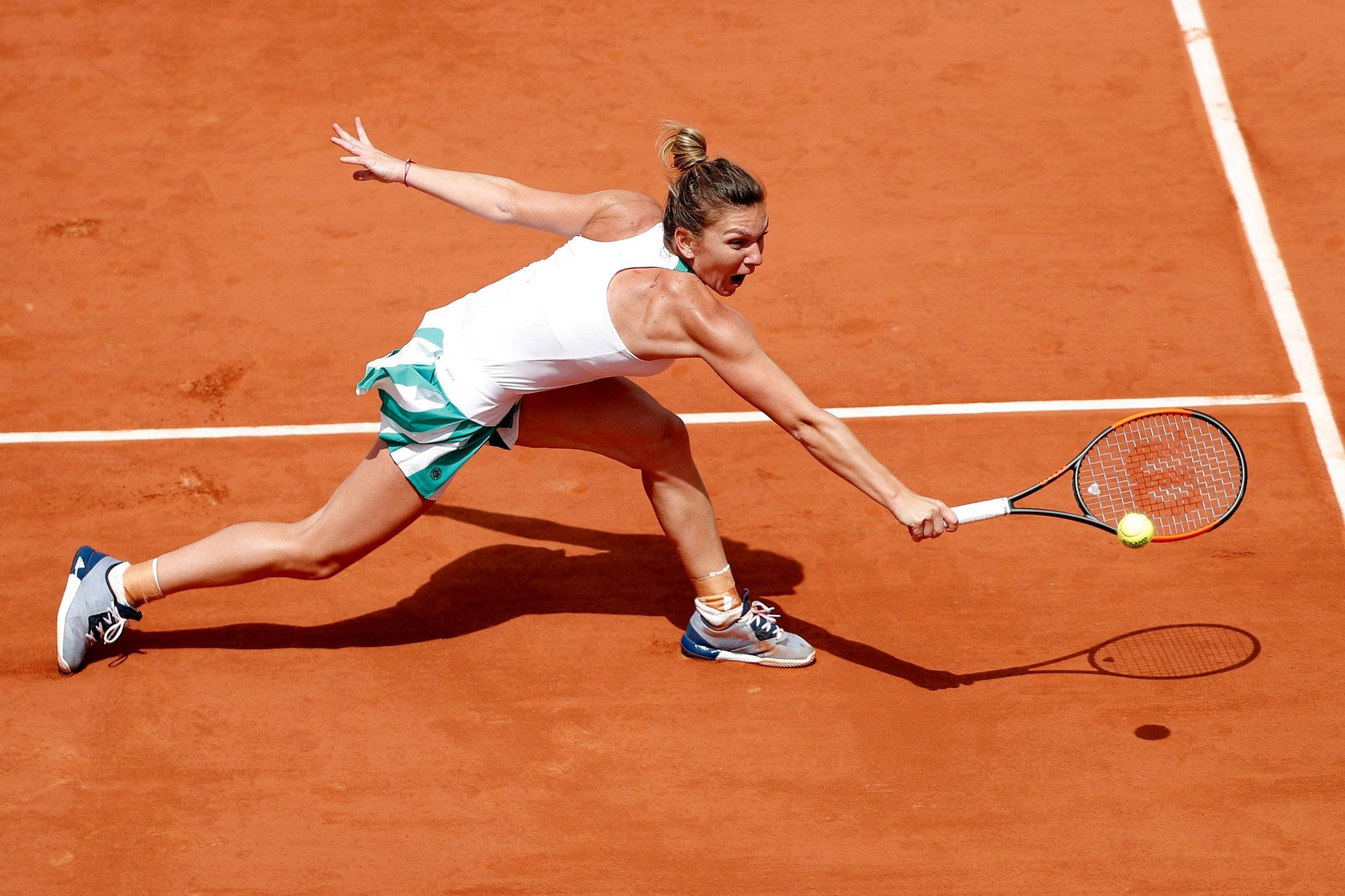Tennis 桌布 壁紙 — 一位女網球選手在紅土球場上向前撲身，在明亮的陽光下以高度專注擊打一個黃色球。