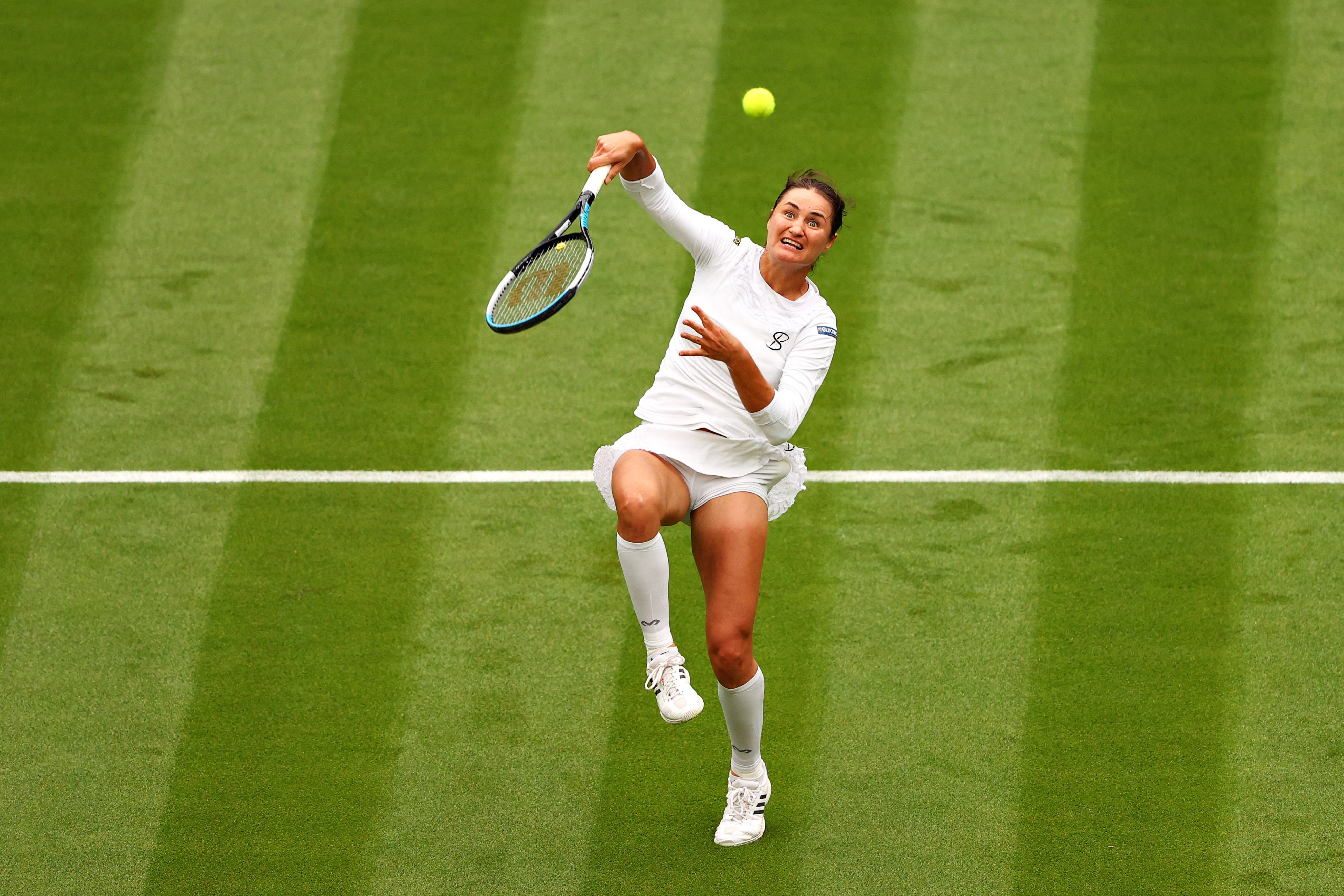 Tennis 桌布 壁紙 — 一位女網球選手跳起擊打一個黃色球，在充滿活力的綠色草地場地上有白色線條。