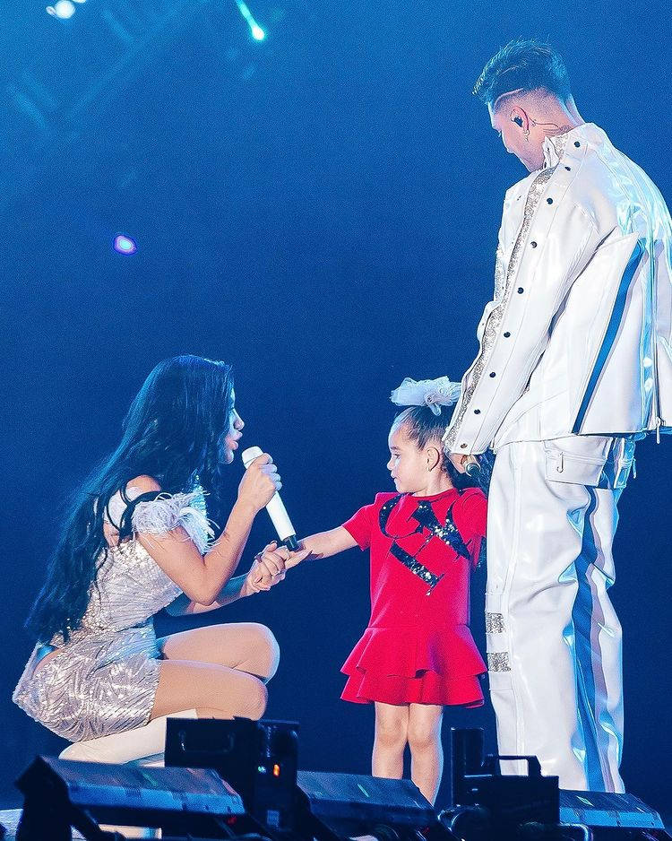 Family 桌布 壁紙 — 一位女表演者跪在舞台上，手持麥克風，與一位穿著紅色連衣裙的小女孩和一位穿著白色西裝的高大男藝人在藍色舞台燈光下互動。
