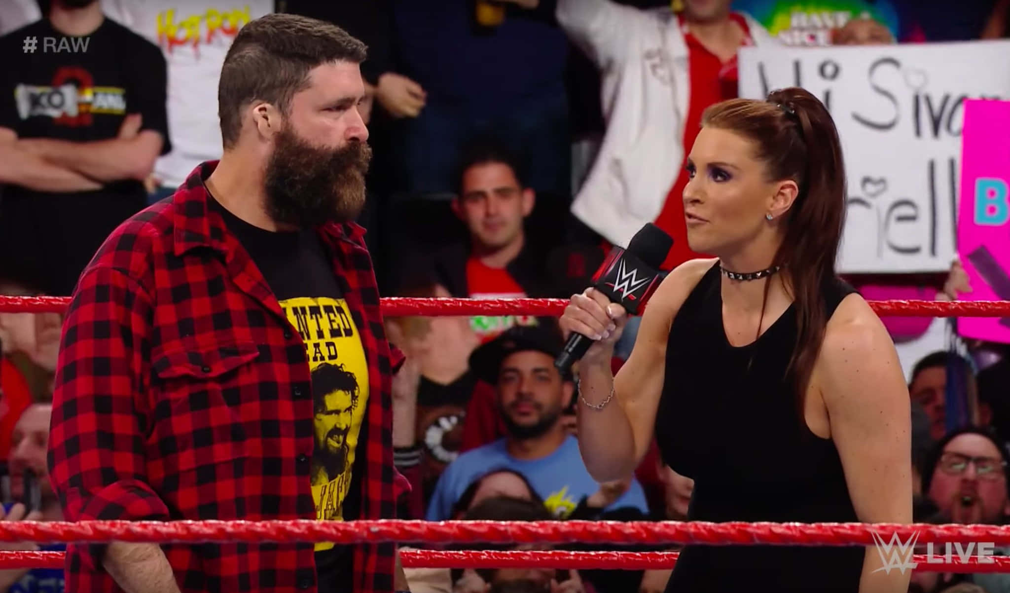 WWE 桌布 壁紙 — 一位女記者拿著麥克風，在摔角場內與一位穿著紅黑格紋襯衫、留鬍子的摔角手對峙，背景有歡呼的粉絲。