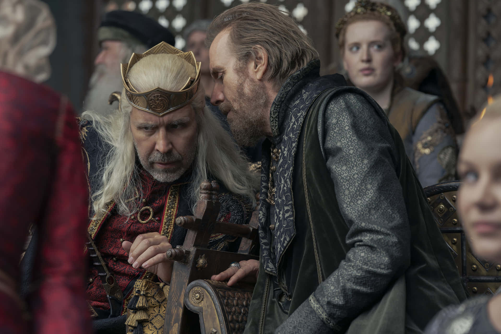 Rhys Ifans 桌布 壁紙 — 一位威嚴的國王戴著金色王冠，留著長長的白髮，坐在王座廳中，一位鬍鬚男急切地俯身在他旁邊說話。