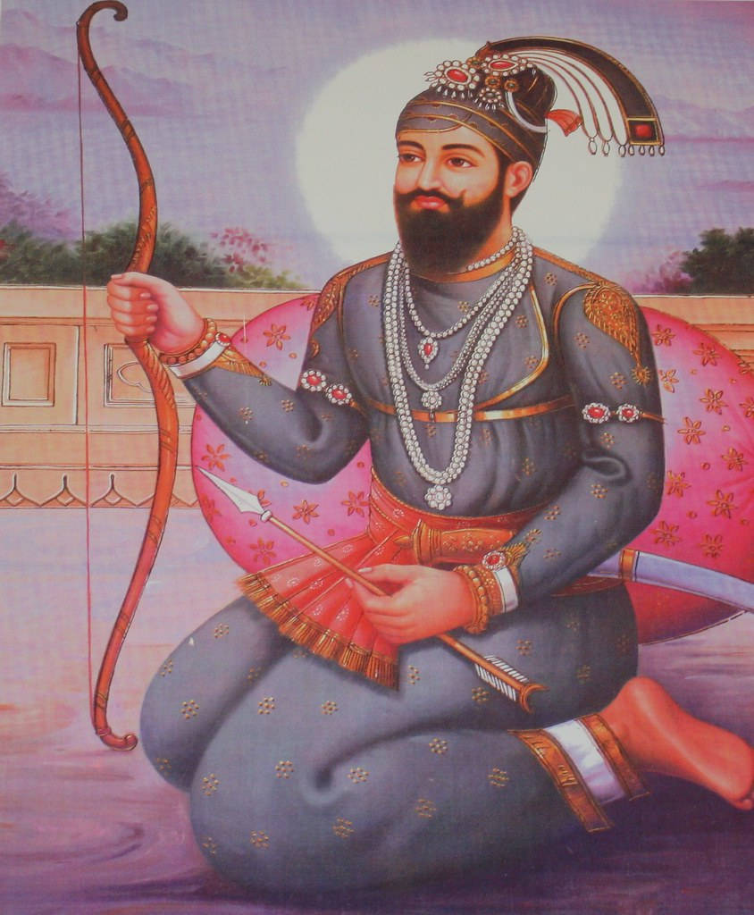 Guru Gobind Singh Ji 桌布 壁紙 — 一位威嚴的鬍鬚男子身穿華麗的藍色和粉紅色服裝，手持弓箭，坐在有軟墊的寶座上，背景是發光的月亮。
