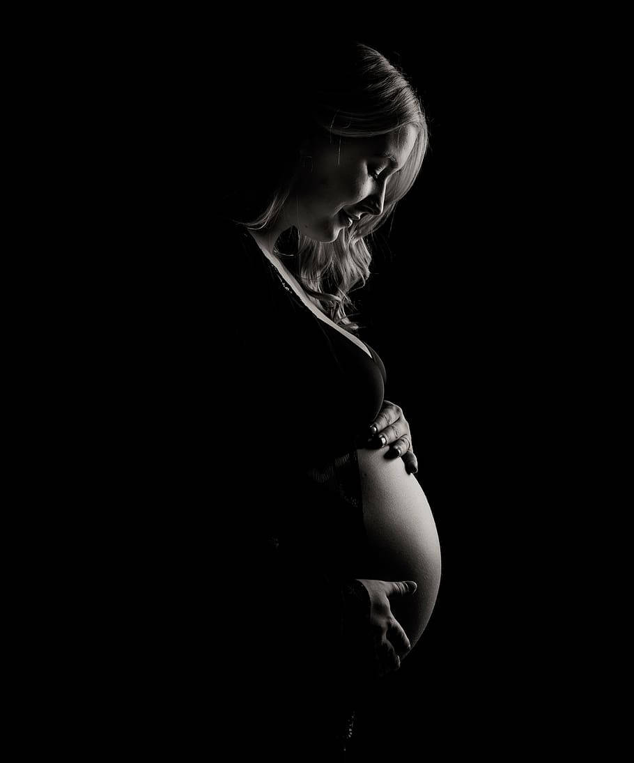 Pregnant Belly 桌布 壁紙 — 一位孕婦以剪影姿態，在純黑背景前輕撫腹部，柔和光線勾勒出她的側面和雙手。
