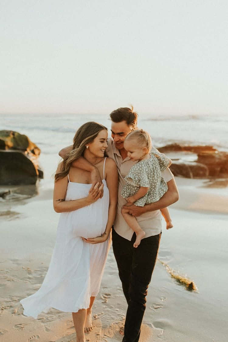 Family Beach 桌布 壁紙 — 一位孕婦和她的伴侶在陽光明媚的海灘上擁抱，抱著他們的幼兒，溫柔的海浪拍打著他們的腳。