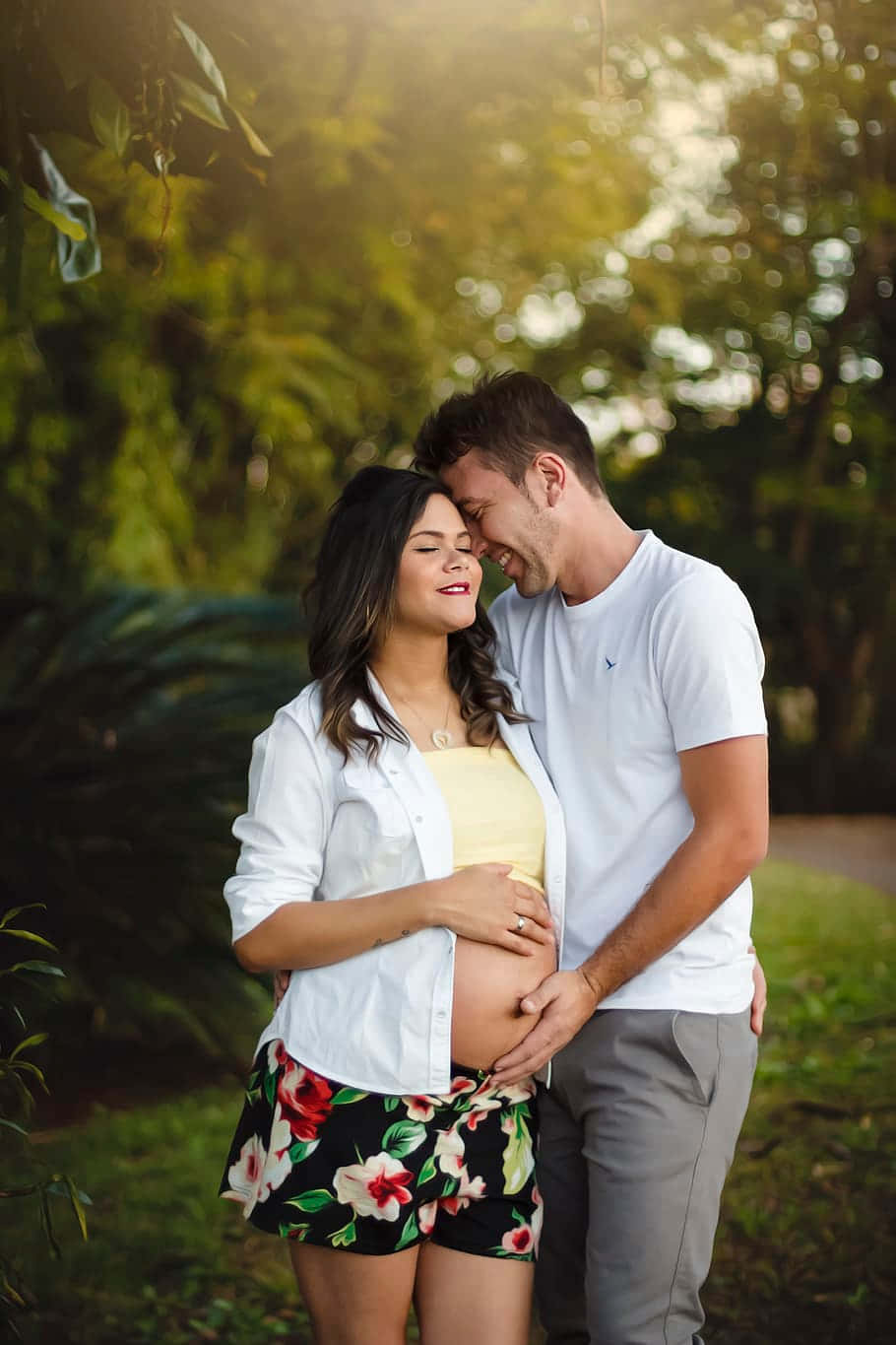 Pregnant 桌布 壁紙 — 一位孕婦和她的伴侶在陽光明媚的花園中溫柔擁抱，額頭相觸，被茂密的綠植環繞。