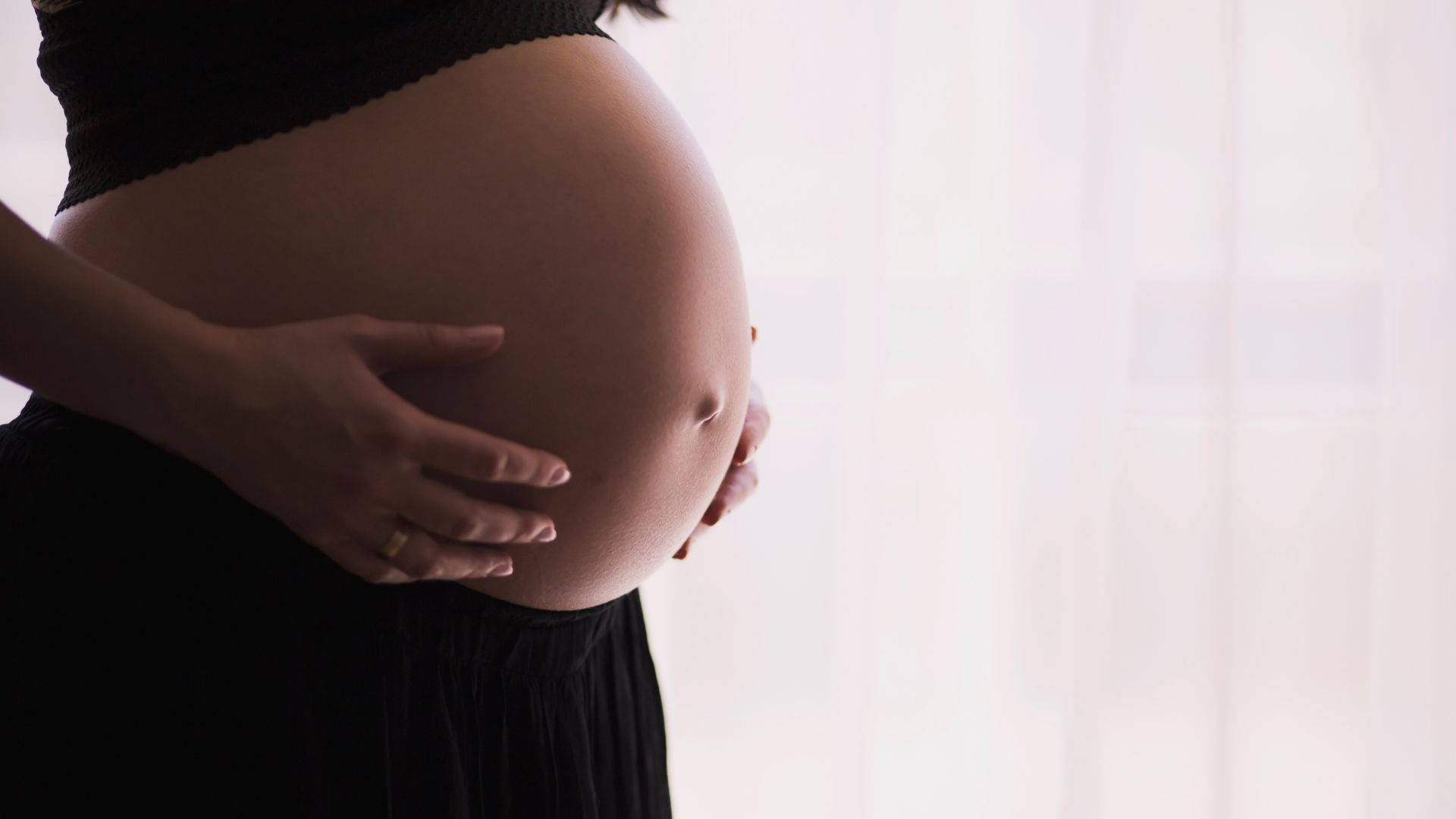 Woman 桌布 壁紙 — 一位孕婦用一隻手輕輕地托著她圓潤的肚子，背景是柔軟的白色窗簾。