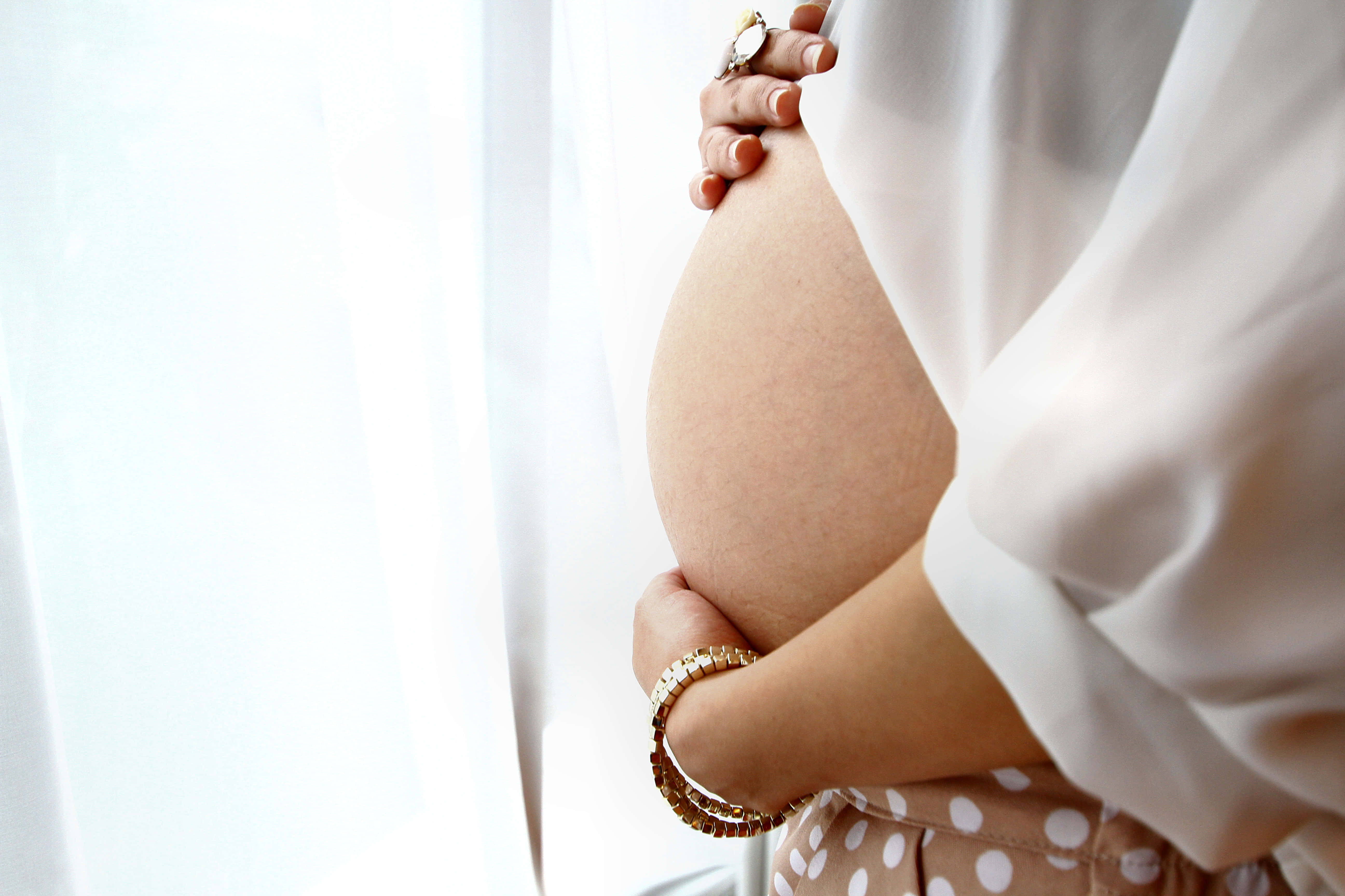 Pregnant 桌布 壁紙 — 一位孕婦用雙手輕輕地抱著肚子，靠近一個有薄窗簾的明亮窗戶。