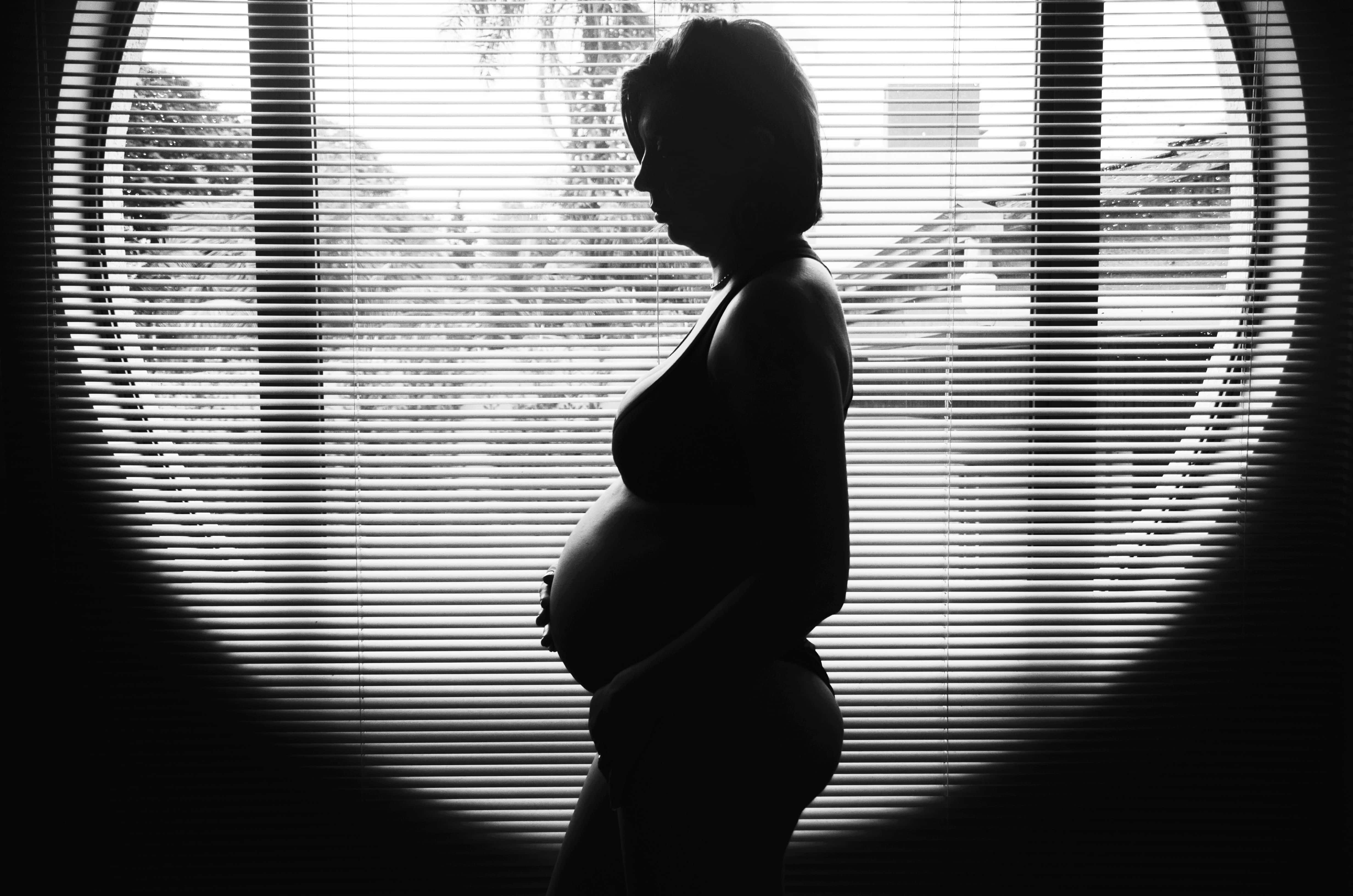 Pregnant 桌布 壁紙 — 一位孕婦的剪影側面站在圓形窗戶前，窗戶有水平百葉窗，雙手捧著腹部，黑白風格。