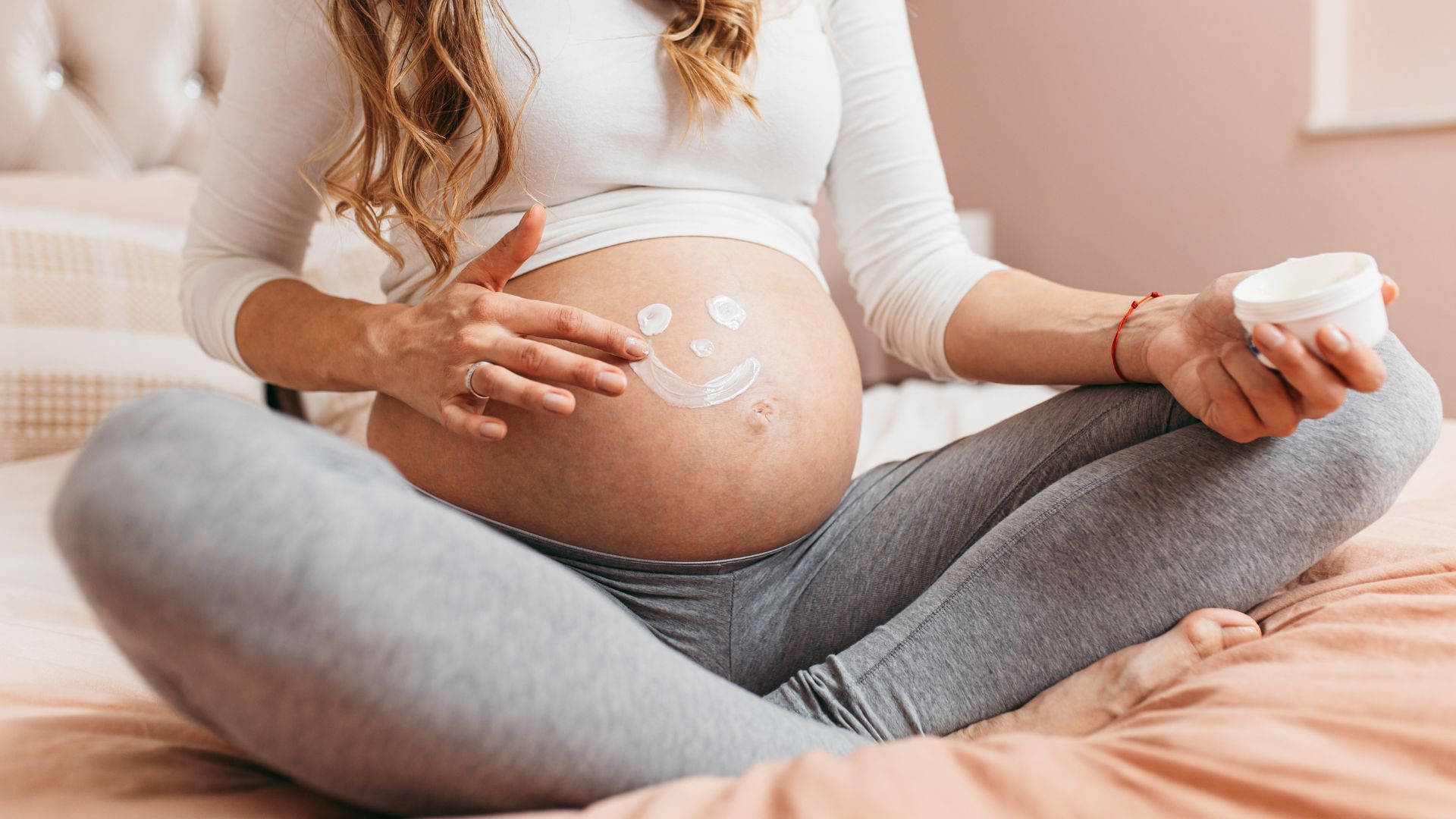Woman 桌布 壁紙 — 一位孕婦盤腿坐在床上，輕輕地在肚子上塗抹乳液，形成笑臉形狀。