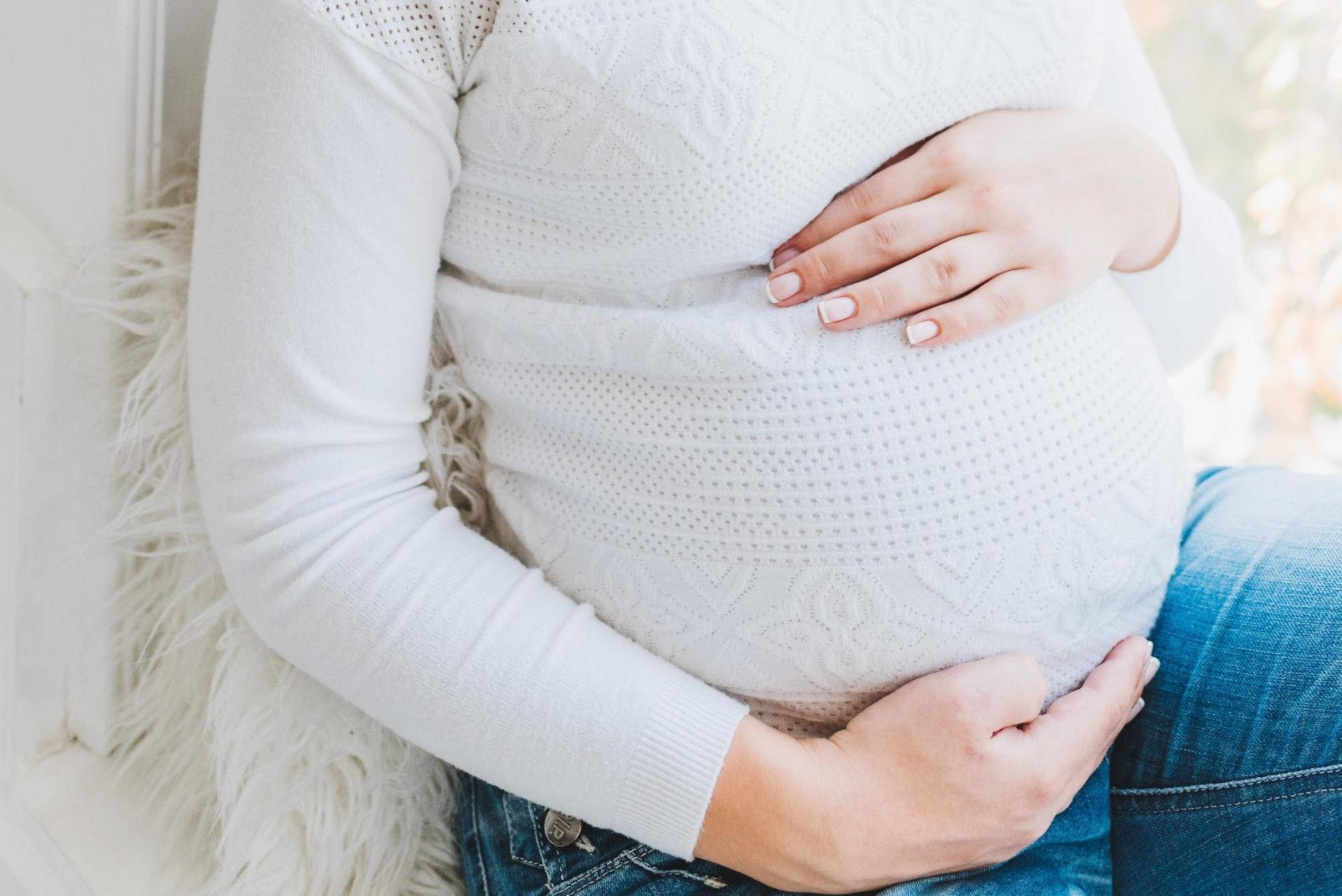 Pregnant 桌布 壁紙 — 一位孕婦穿著柔軟的白色毛衣，輕輕地抱著肚子，坐在一個蓬鬆的白色坐墊上。